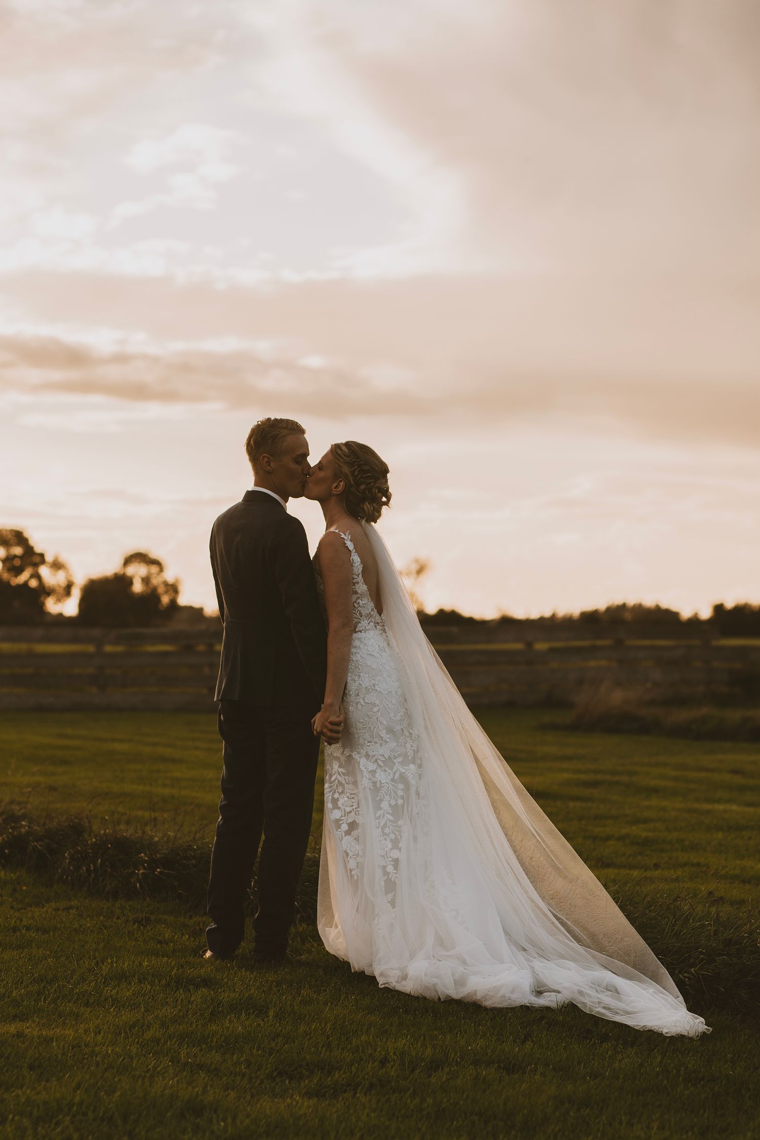 A silhouetted couple shares an intimate moment at sunset with a flowing wedding dress in view.