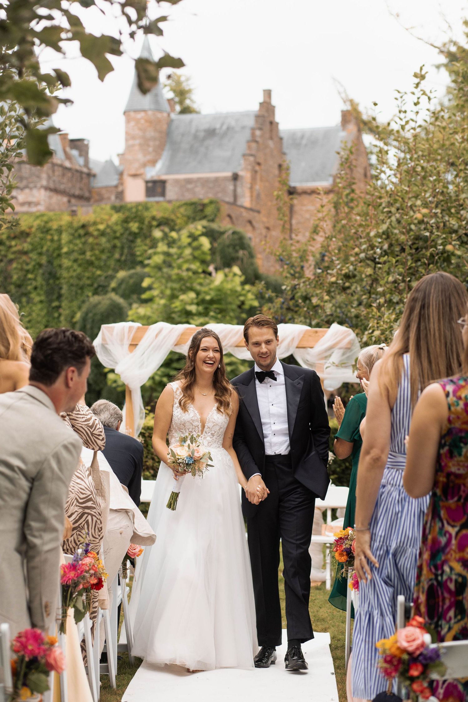 Bride and groom walk down outdoor aisle at castle wedding ceremony, guests seated with colorful floral arrangements.