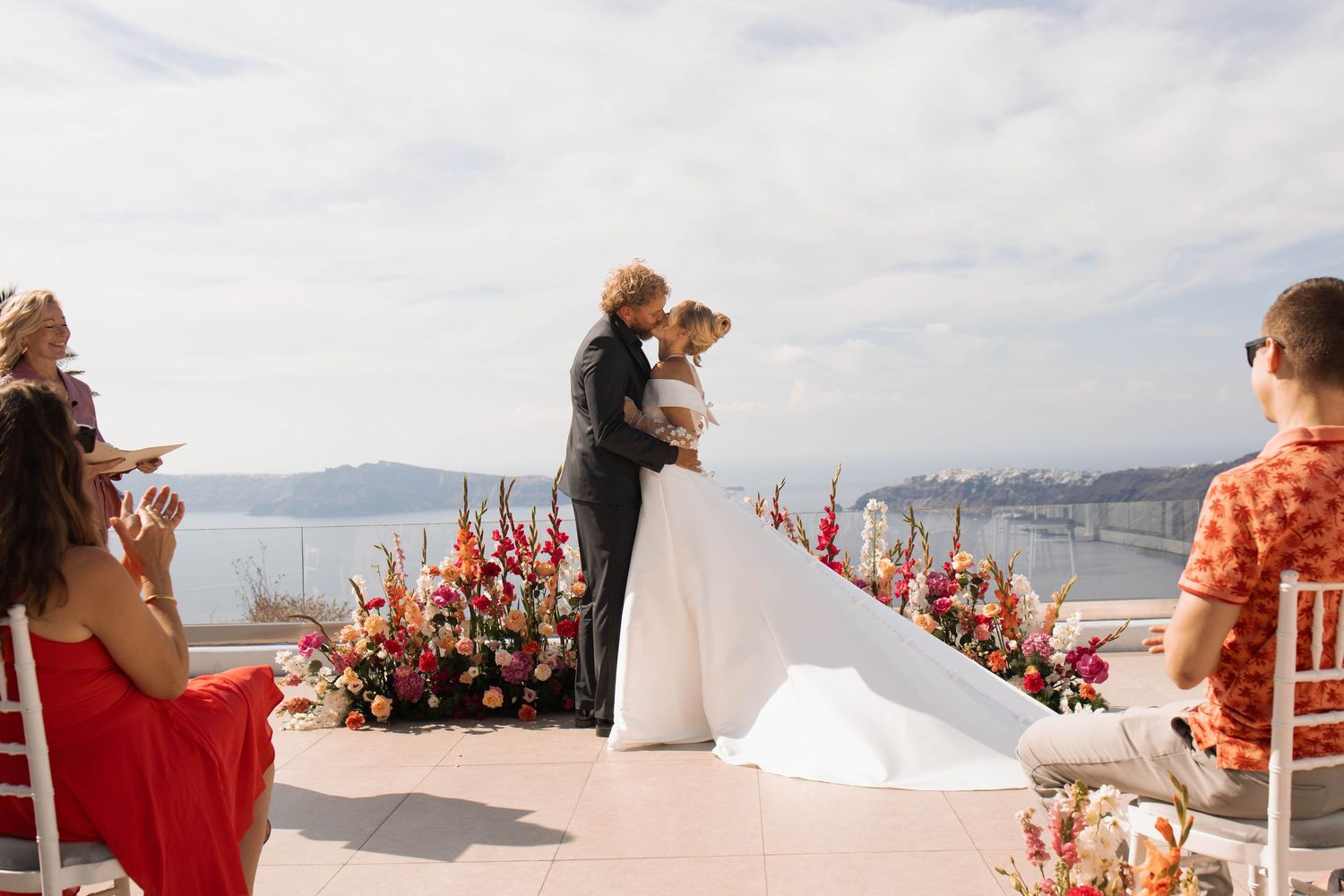 Couple shares a kiss during an outdoor seaside wedding ceremony with colorful flower arrangements and guests watching overlooking the water.