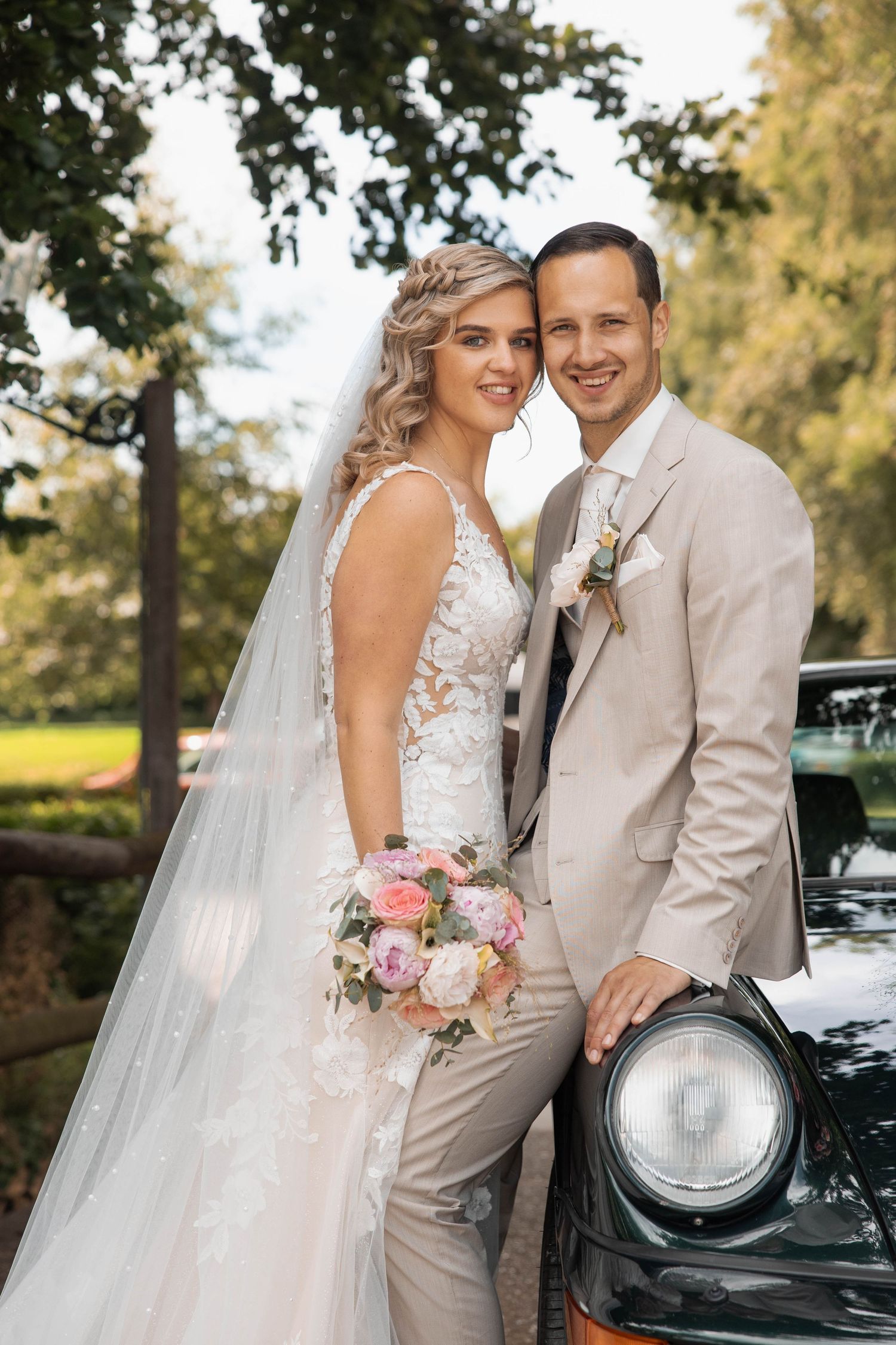 Bride and groom posing beside a vintage car outdoors, bride holding pink bouquet in lace gown with veil.