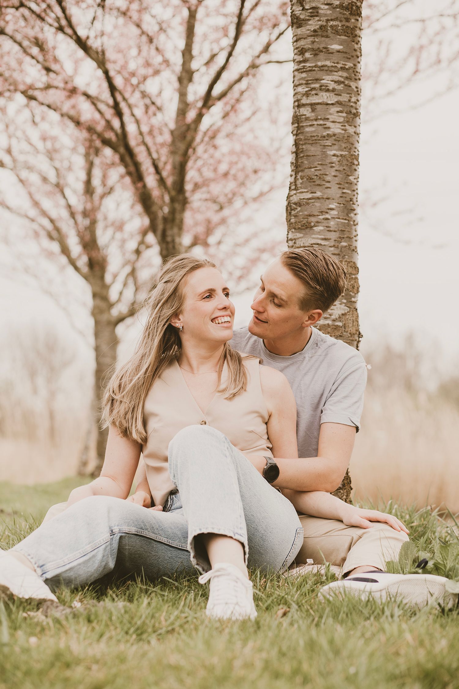 A couple relaxes together on grass beneath blooming spring trees in a romantic outdoor photoshoot.