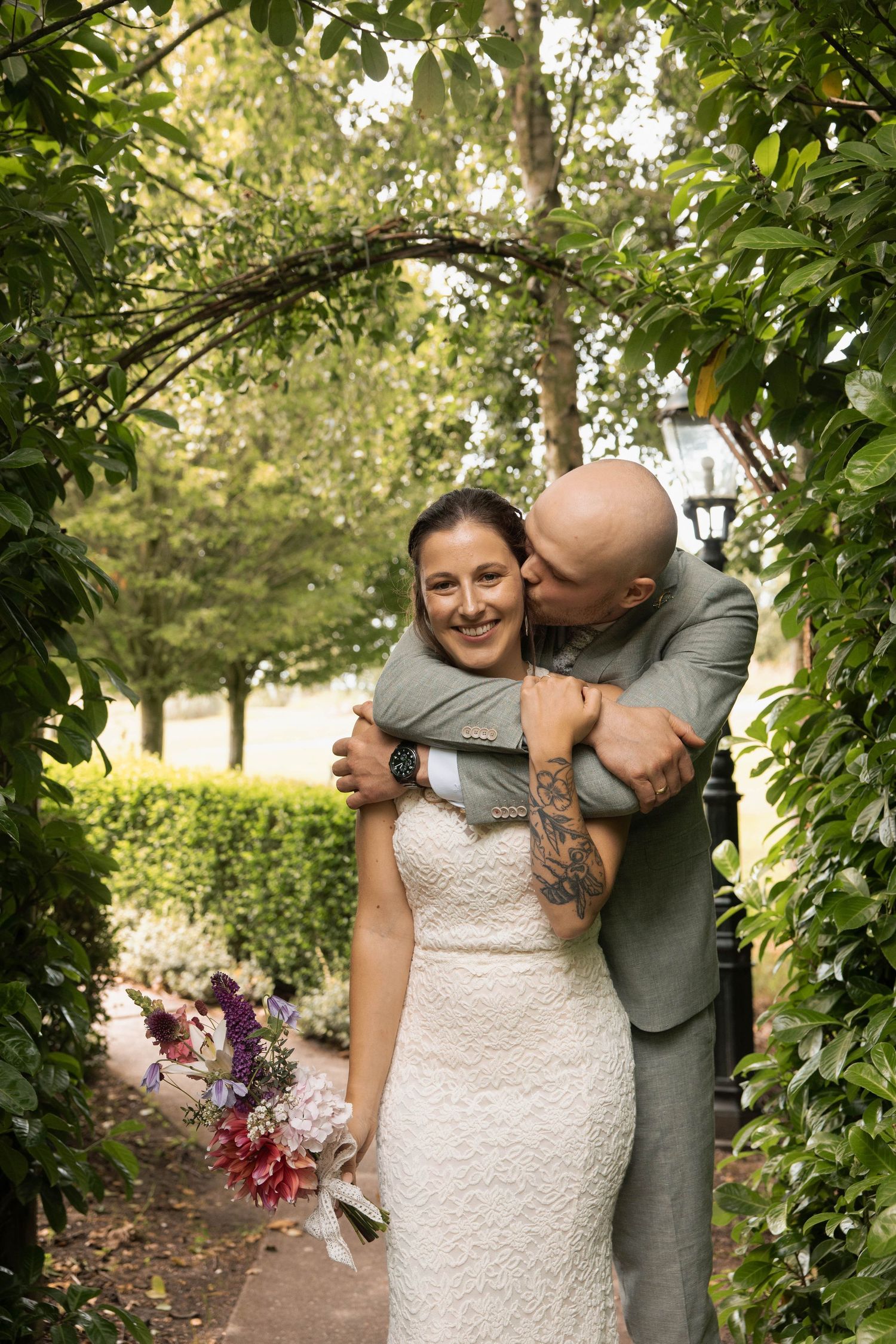 Groom with tattoos embraces bride in white lace dress holding colorful bouquet under garden arch.
