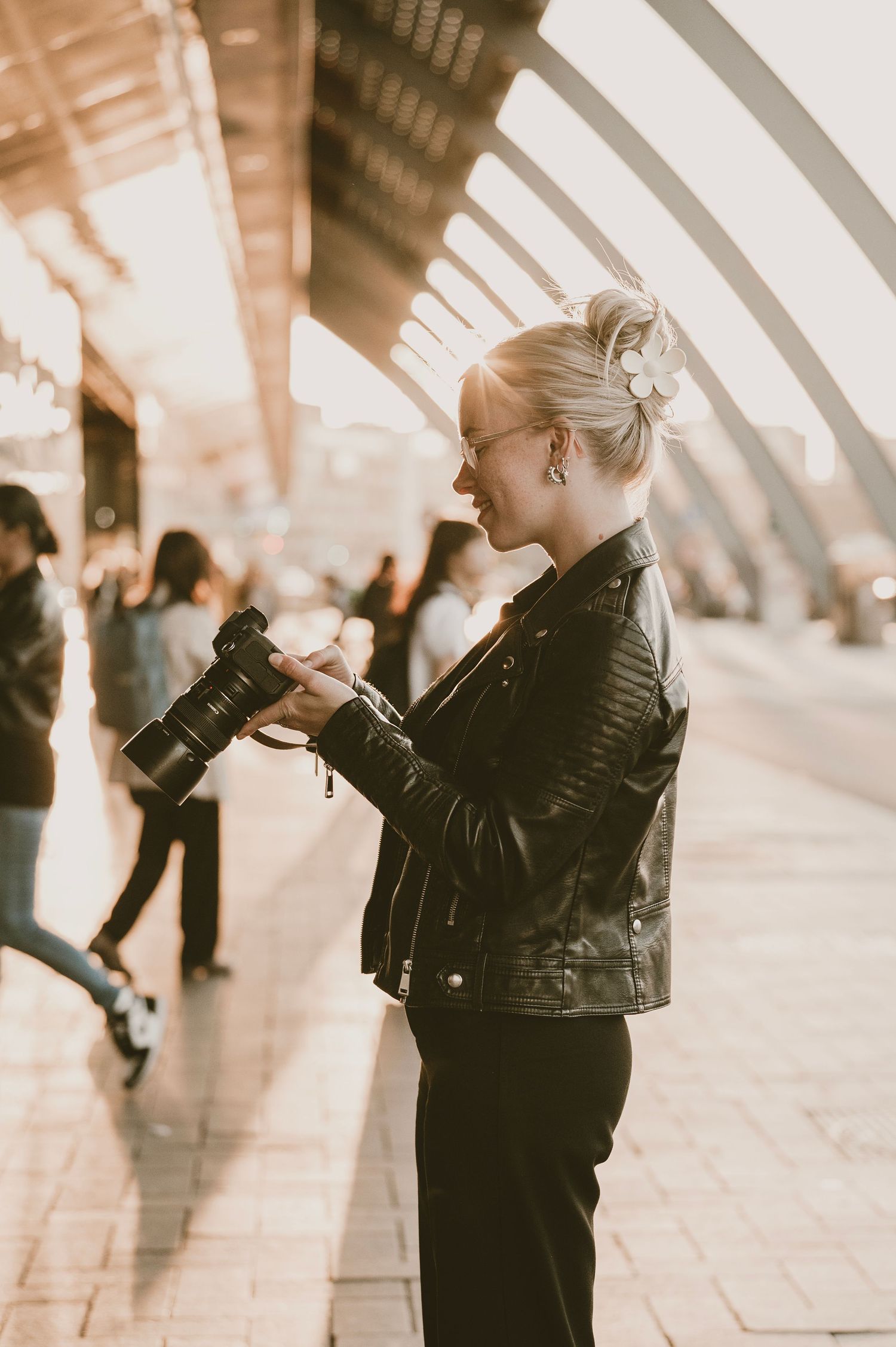 A person in a black leather jacket stands in a sunlit urban setting with geometric architecture in the background.