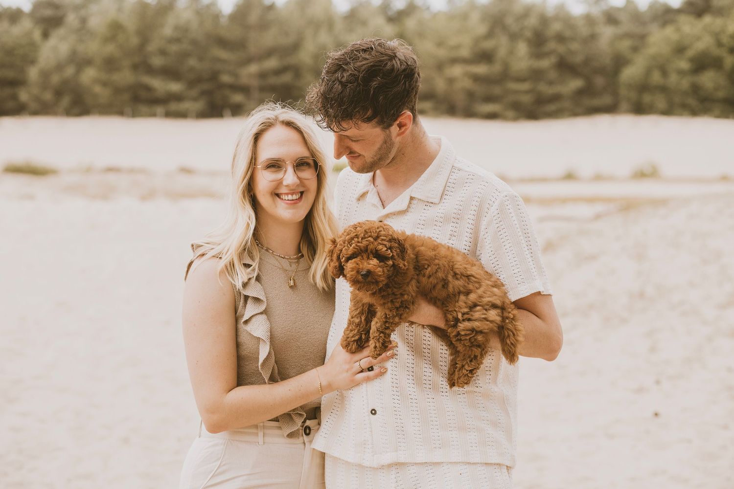 Couple embraces on a sandy beach while holding their brown dog during a warm sunset photoshoot.