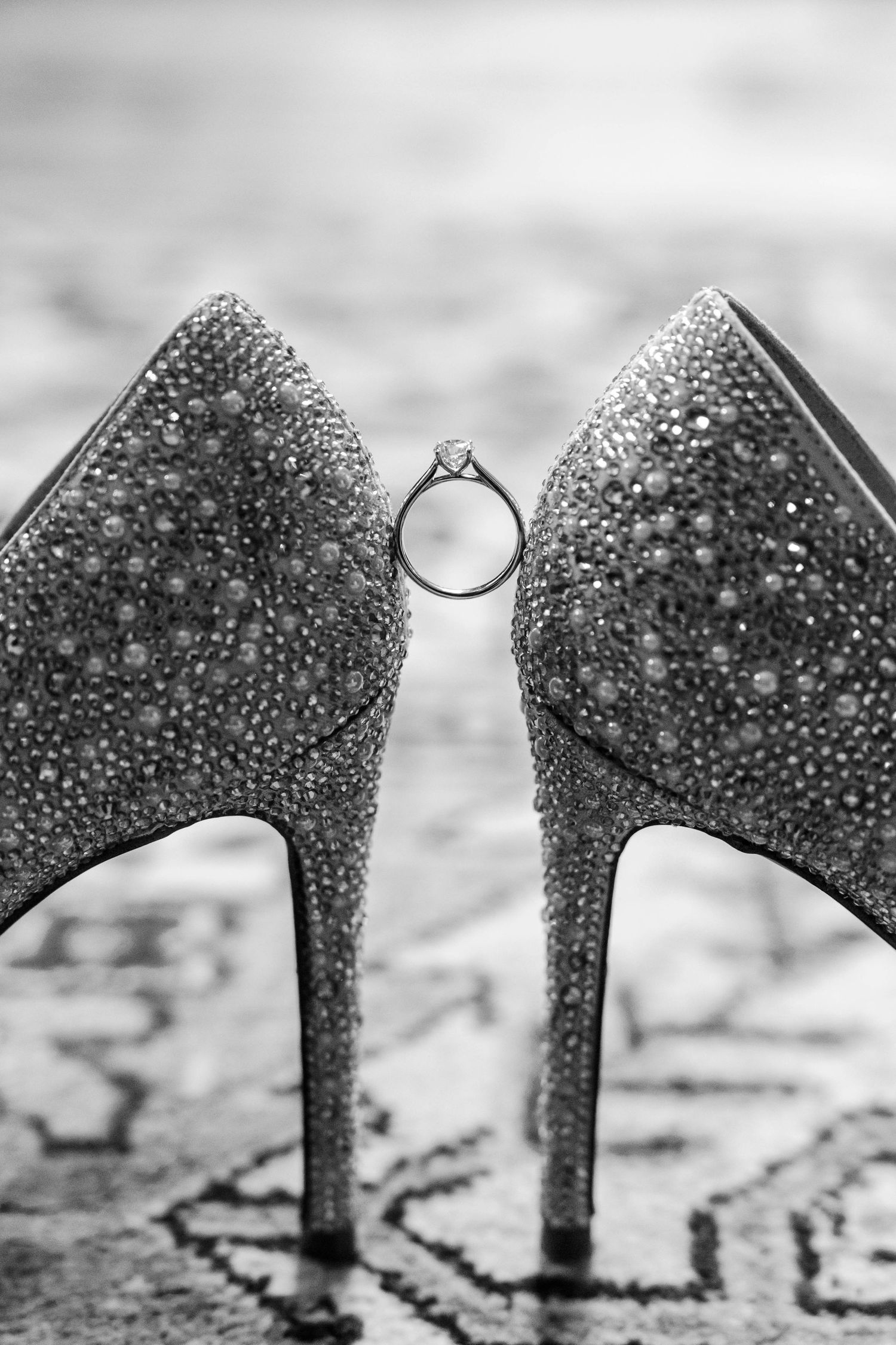 Glittering crystal-encrusted high heel shoes with a diamond engagement ring balanced between them in black and white.