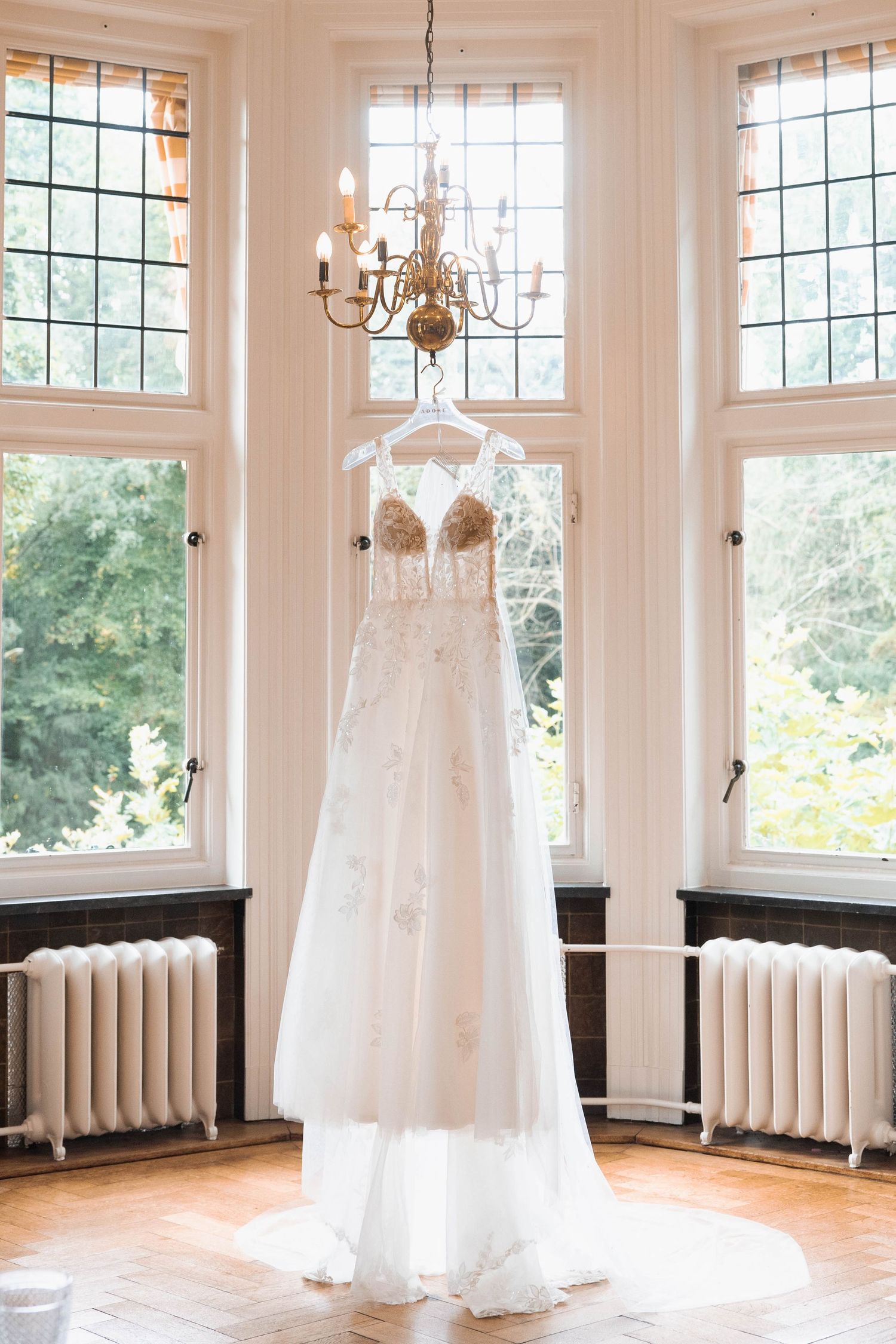 Elegant beaded wedding dress hanging from a chandelier in a bright room with leaded glass windows and white radiators.