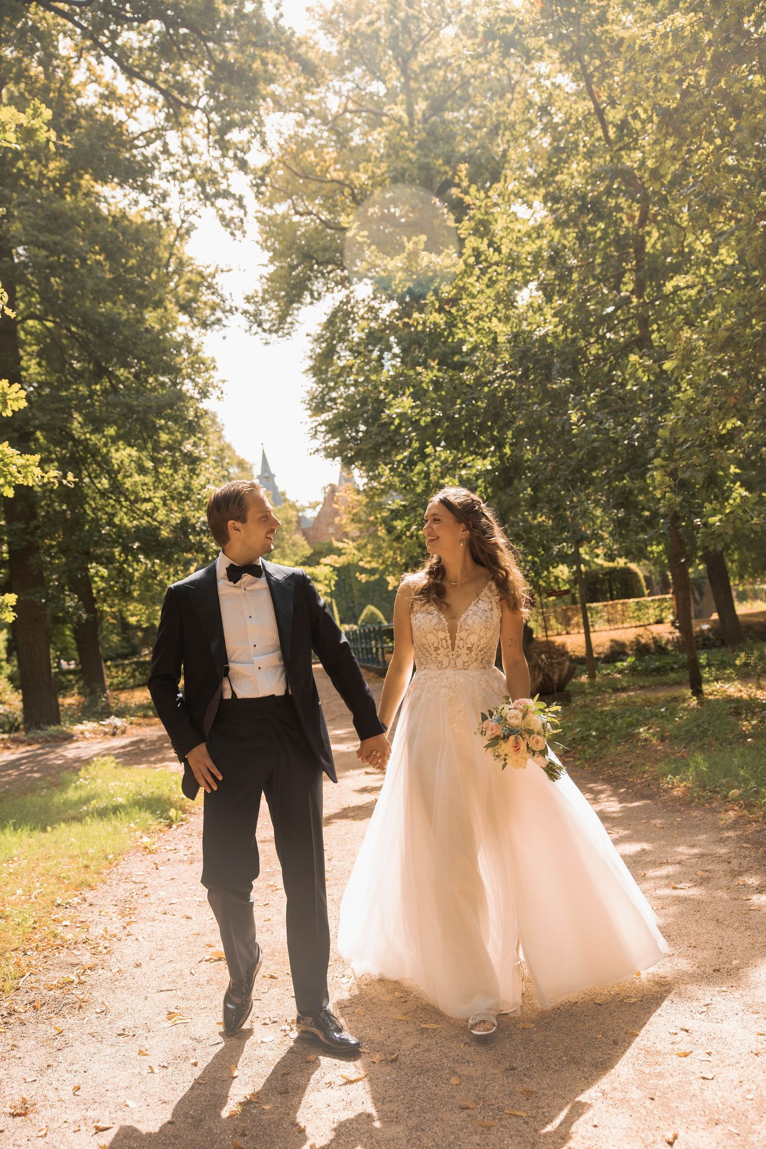 Bride and groom walking hand in hand through a sunlit park path, smiling at each other on their wedding day.