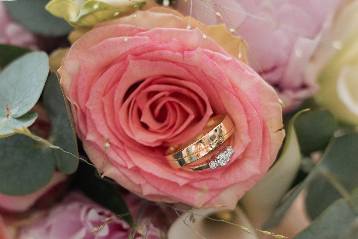 Gold wedding rings with diamonds resting inside a pink rose bloom surrounded by a bridal bouquet.