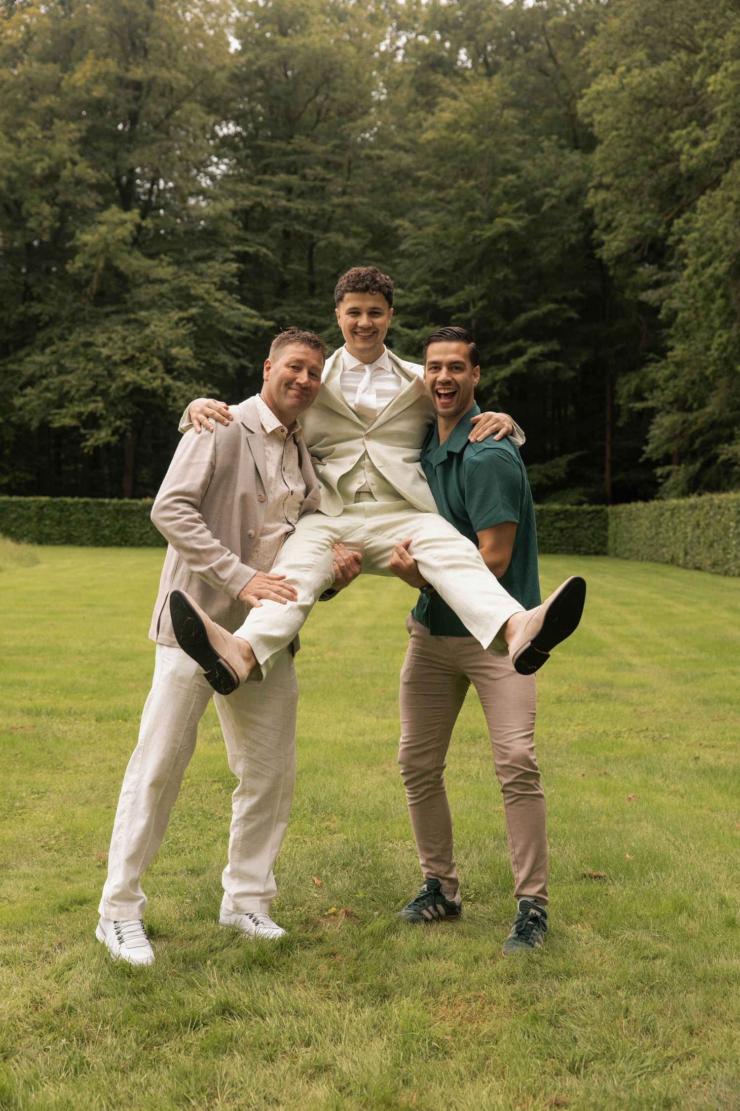 Three men celebrate outdoors on grass, two friends lifting a groom in wedding attire, with lush green trees behind them.