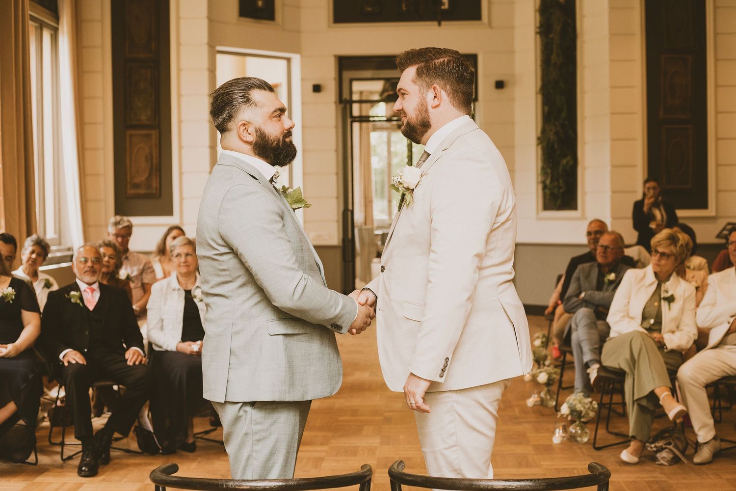 Two people in light colored suits exchange vows during an indoor wedding ceremony.