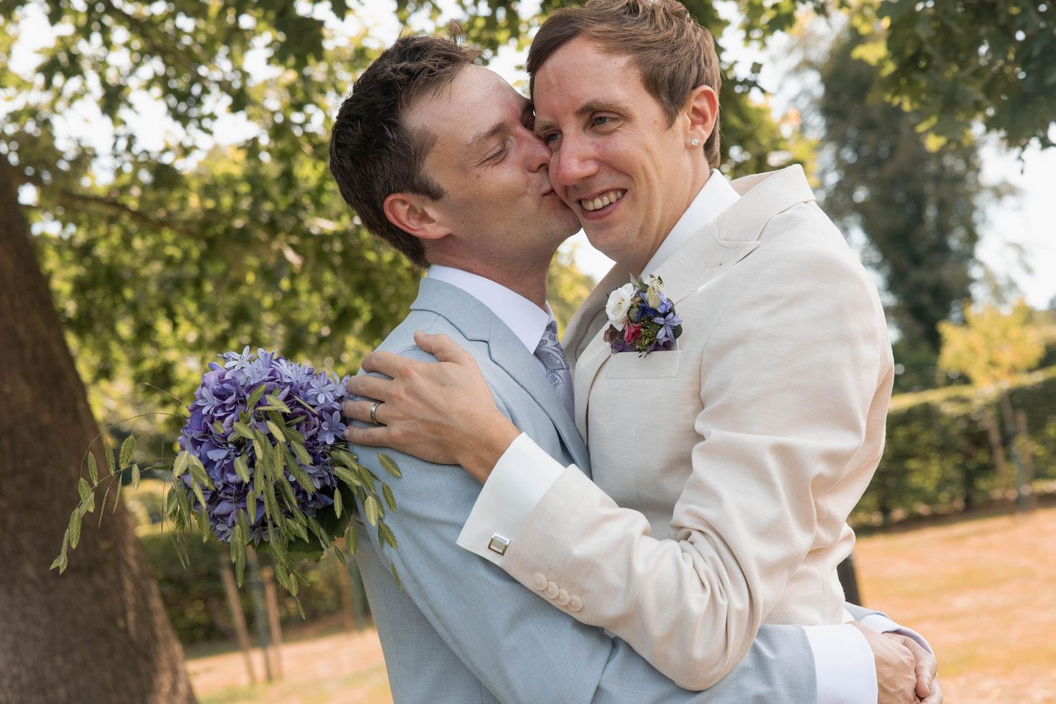 Two grooms in white suits share a romantic kiss outdoors, holding purple flowers at their wedding ceremony.