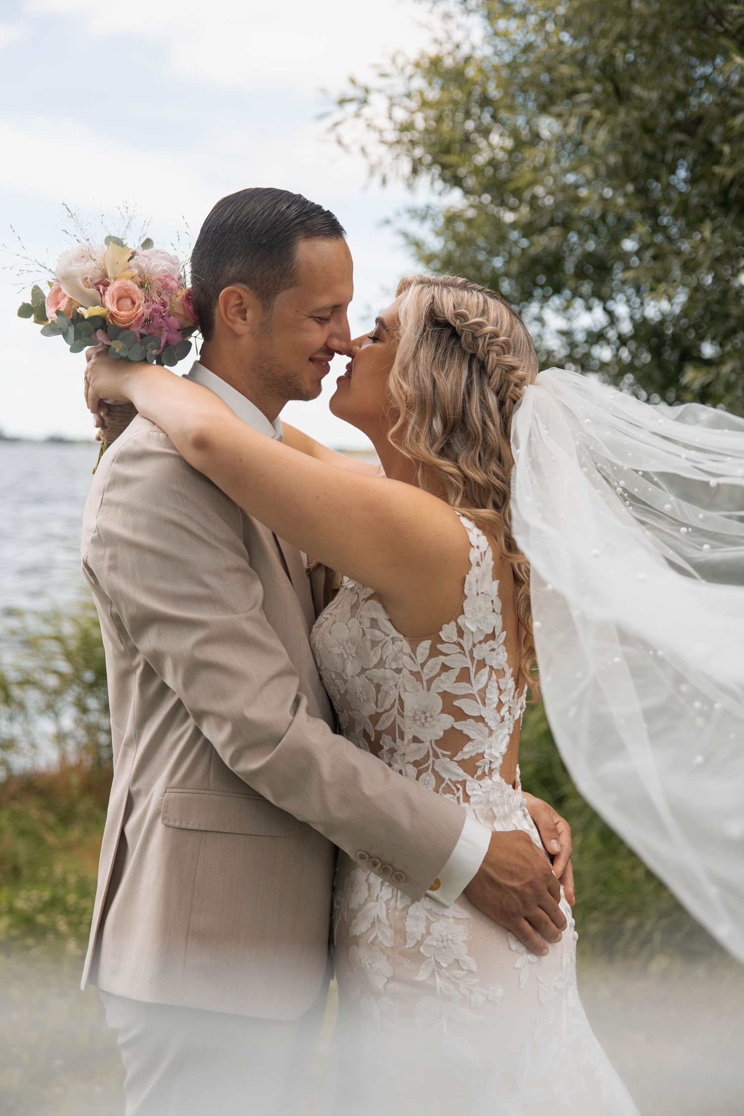 A bride and groom share a romantic almost-kiss outdoors, her lace gown and veil flowing in the breeze near a lakeside.