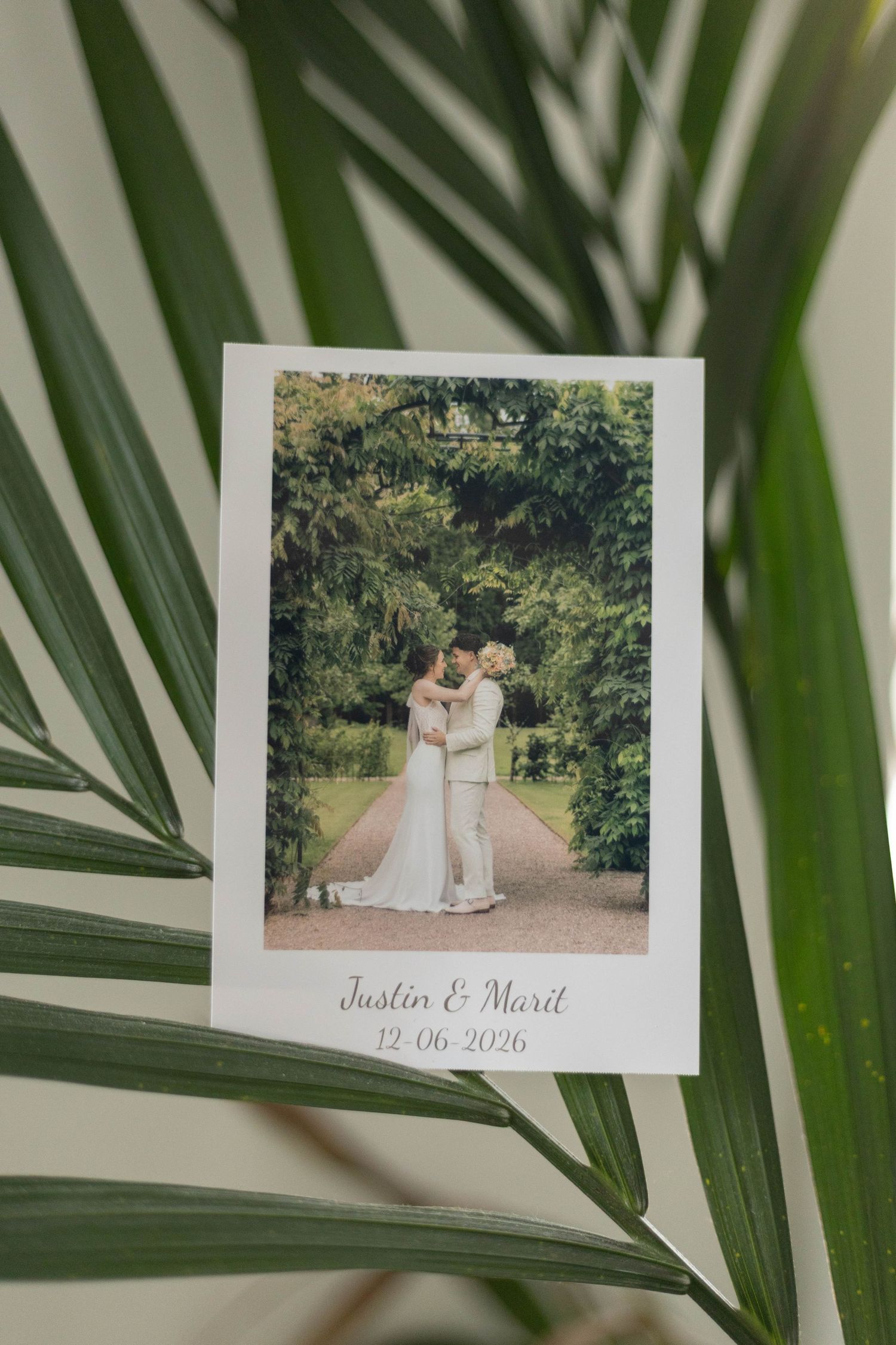 Polaroid wedding photo of Justin and Marit dated 12-06-2026, couple embracing on a garden path, displayed on palm leaves.