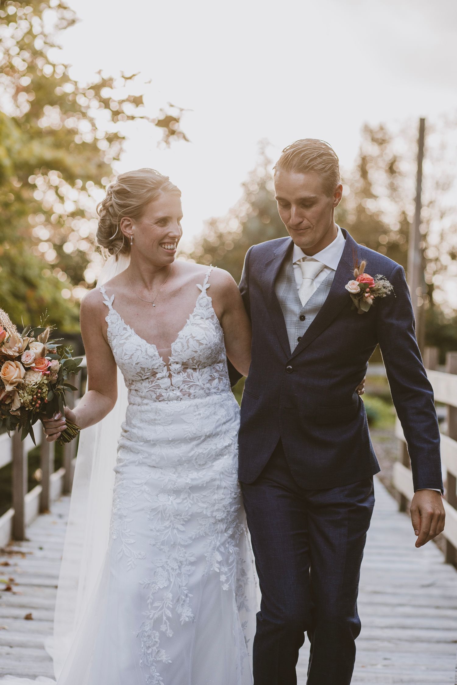 A couple in wedding attire walking and laughing together along a tree-lined path during golden hour.