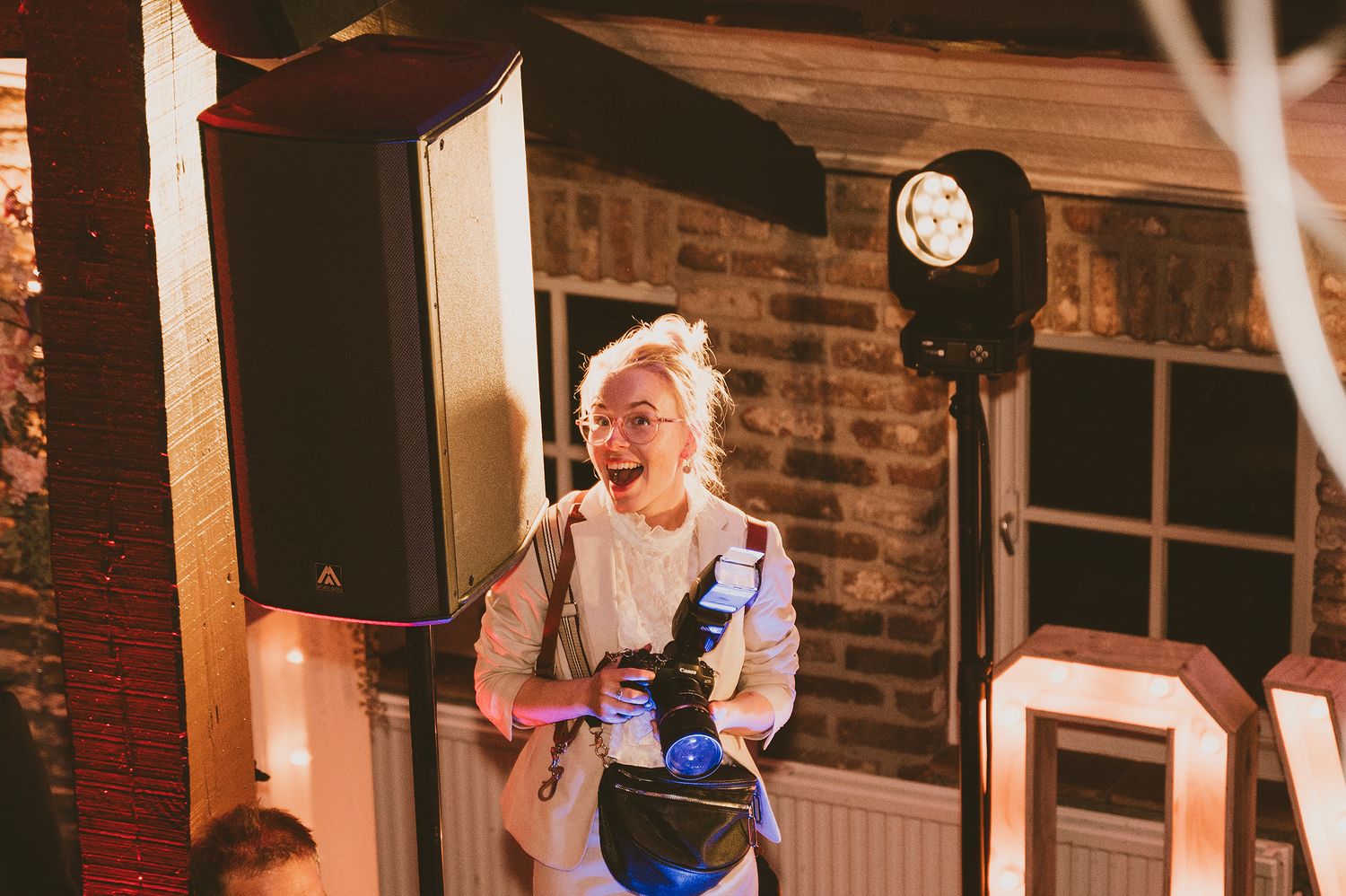 A smiling blonde woman holding a camera at an outdoor evening event with speakers and lights.