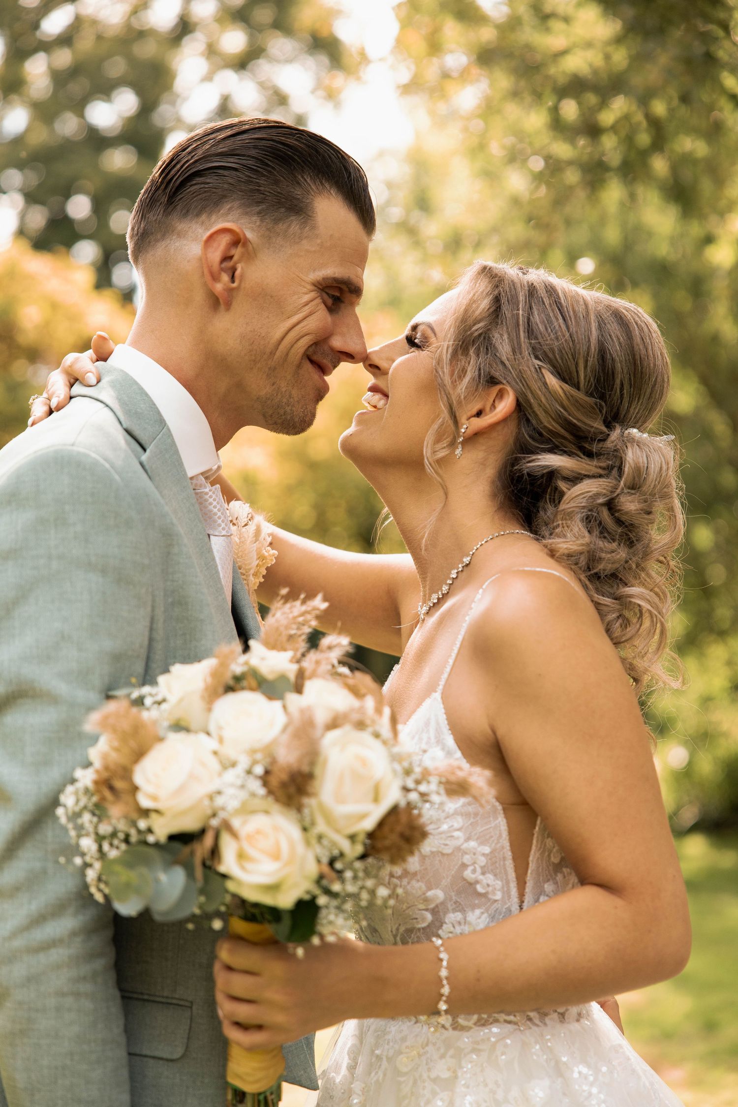 A bride and groom share a romantic moment outdoors, with the bride holding a white rose bouquet in a lace gown.