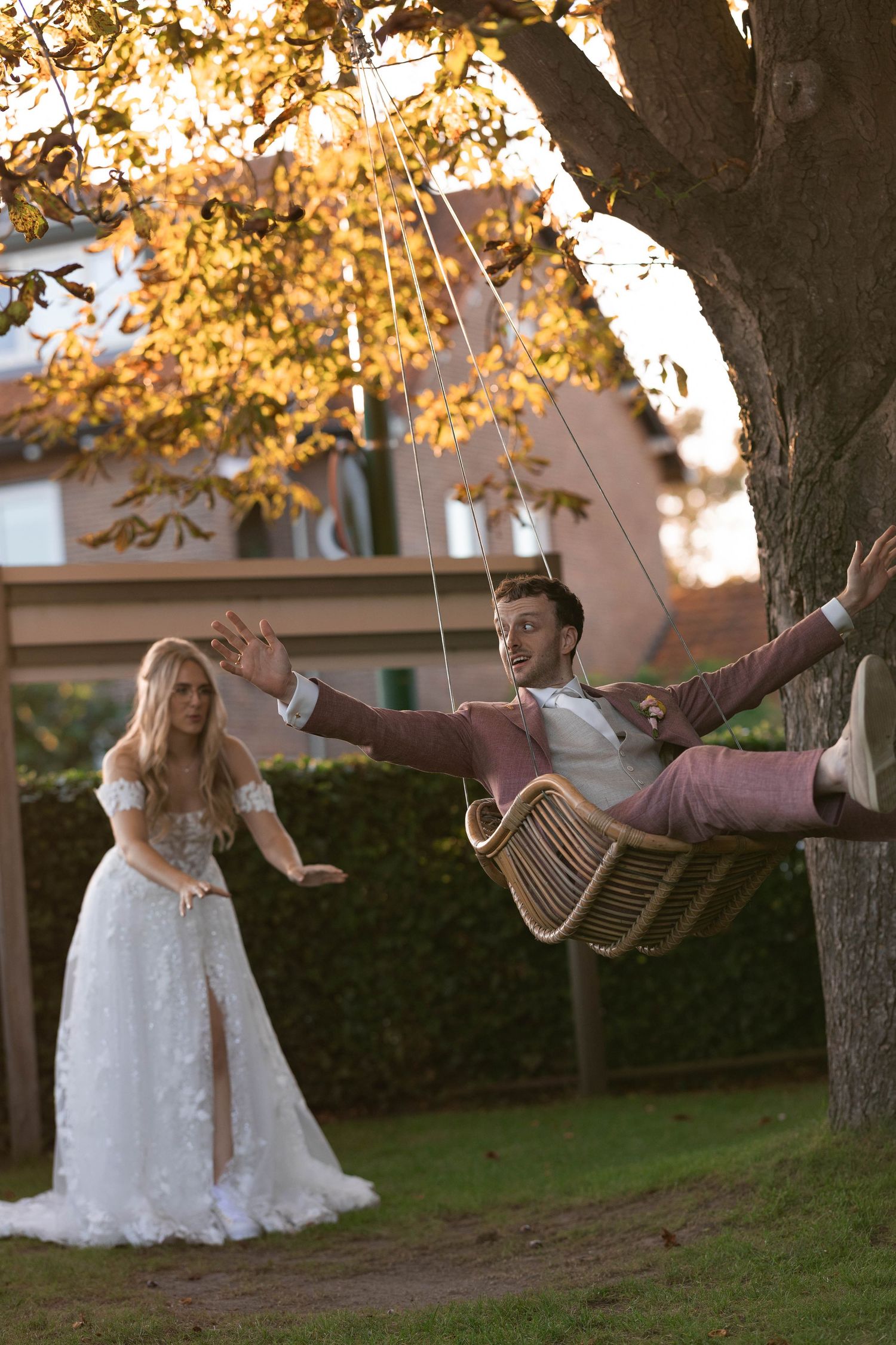 Bride in a white lace gown watches smiling groom in a pink suit lean back on a wicker tree swing in autumn.