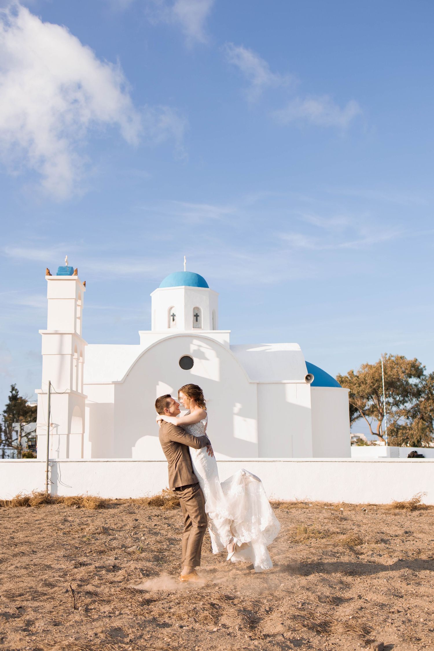 Groom lifts and embraces his bride in a flowing white gown in front of a whitewashed Greek chapel with blue domes under a bright sky.