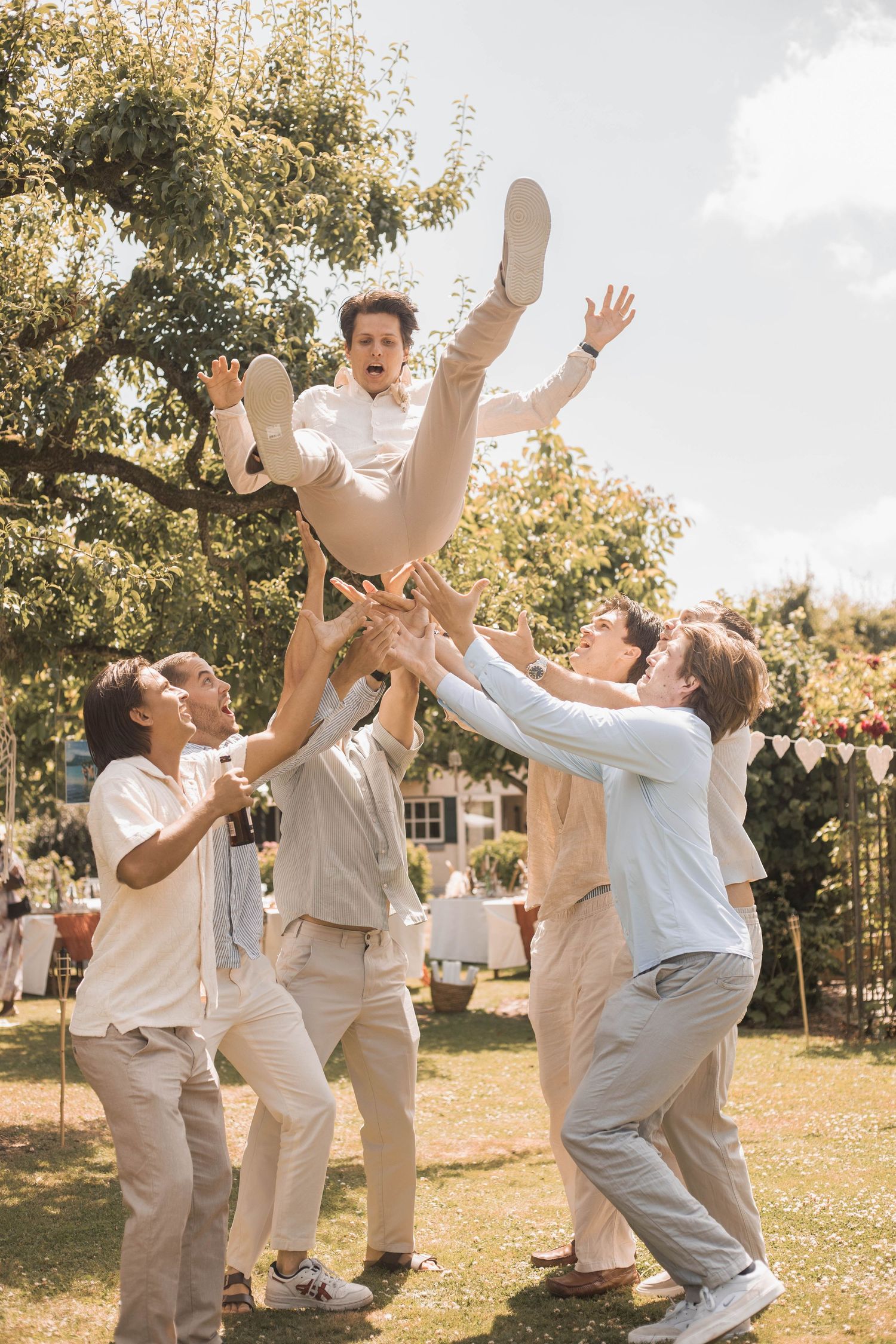 Groomsmen tossing groom in the air during outdoor wedding celebration in garden.