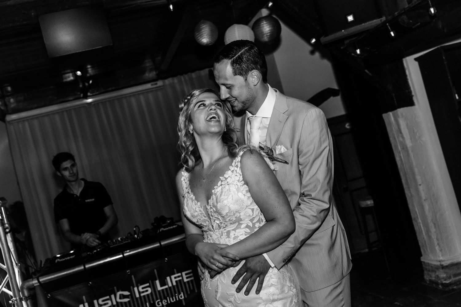 Black and white photo of a bride and groom sharing their first dance at a wedding reception with a DJ in the background.