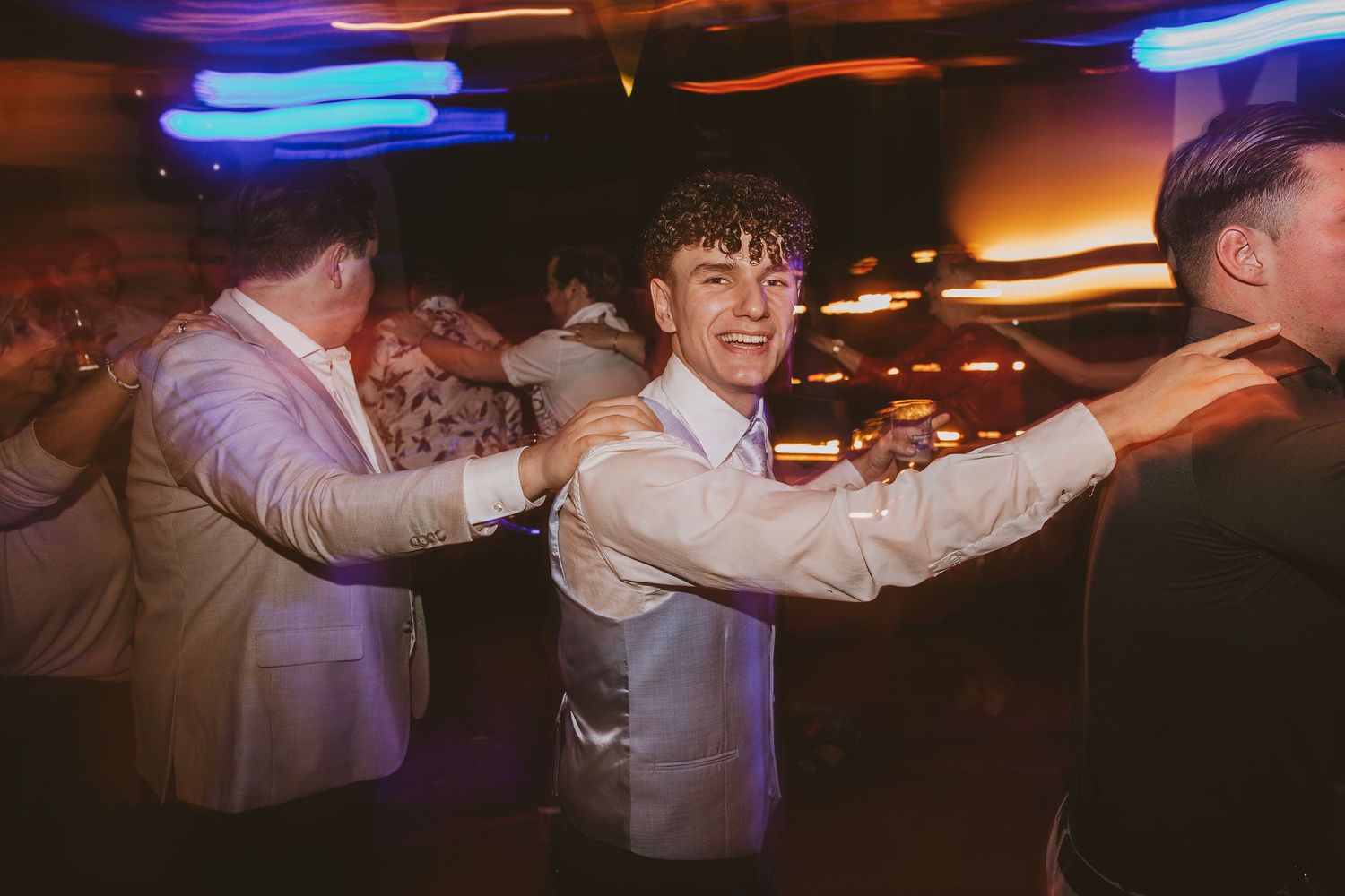 Wedding guests form a line with arms around each other's shoulders during a reception celebration.