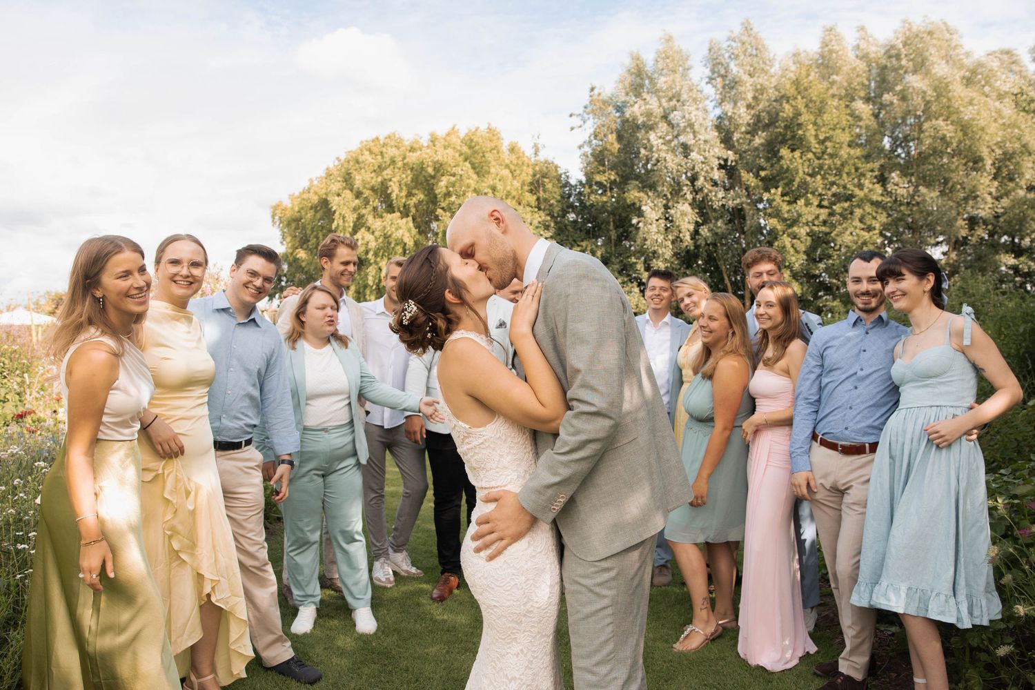Bride and groom share a kiss surrounded by smiling wedding guests on a lush green lawn.