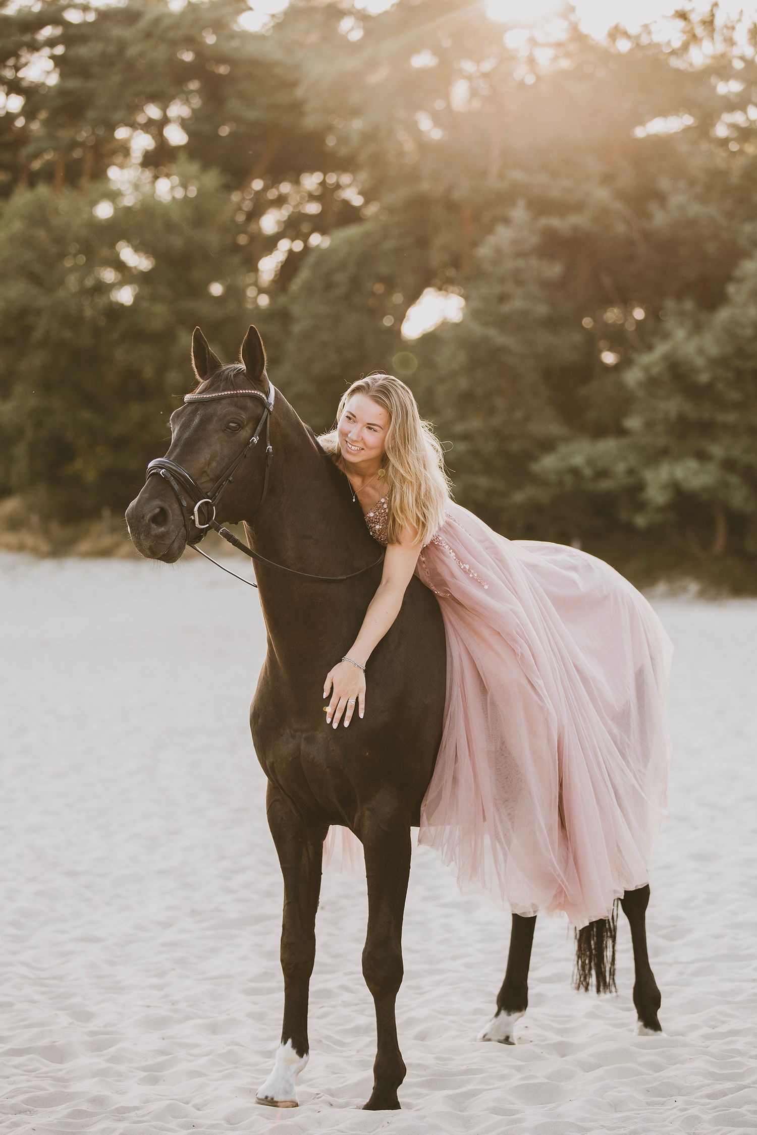 Woman in flowing pink dress poses with black horse on sandy beach at sunset.