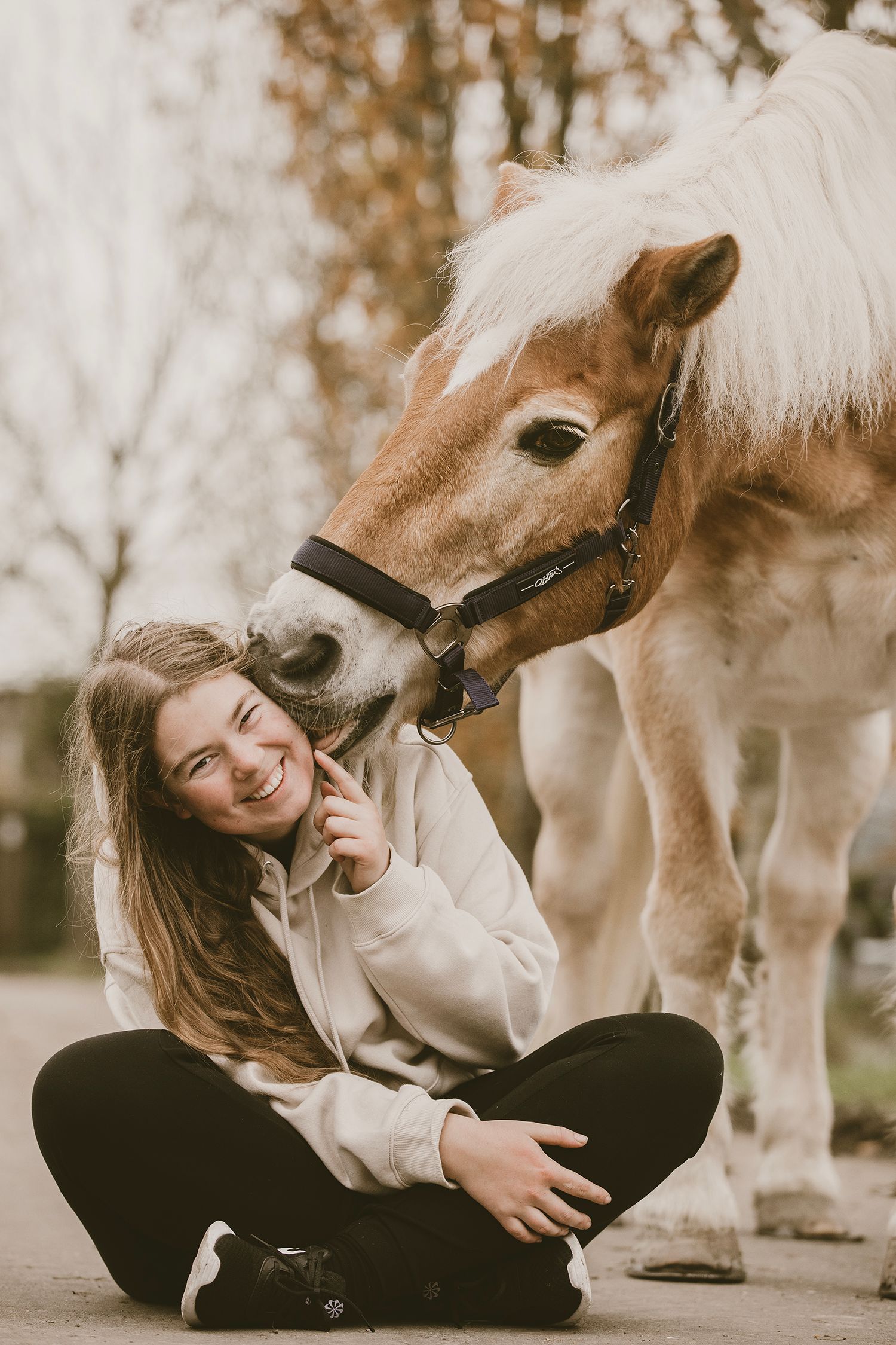 A gentle moment between a cream-colored horse and someone sitting cross-legged on the ground in casual attire.
