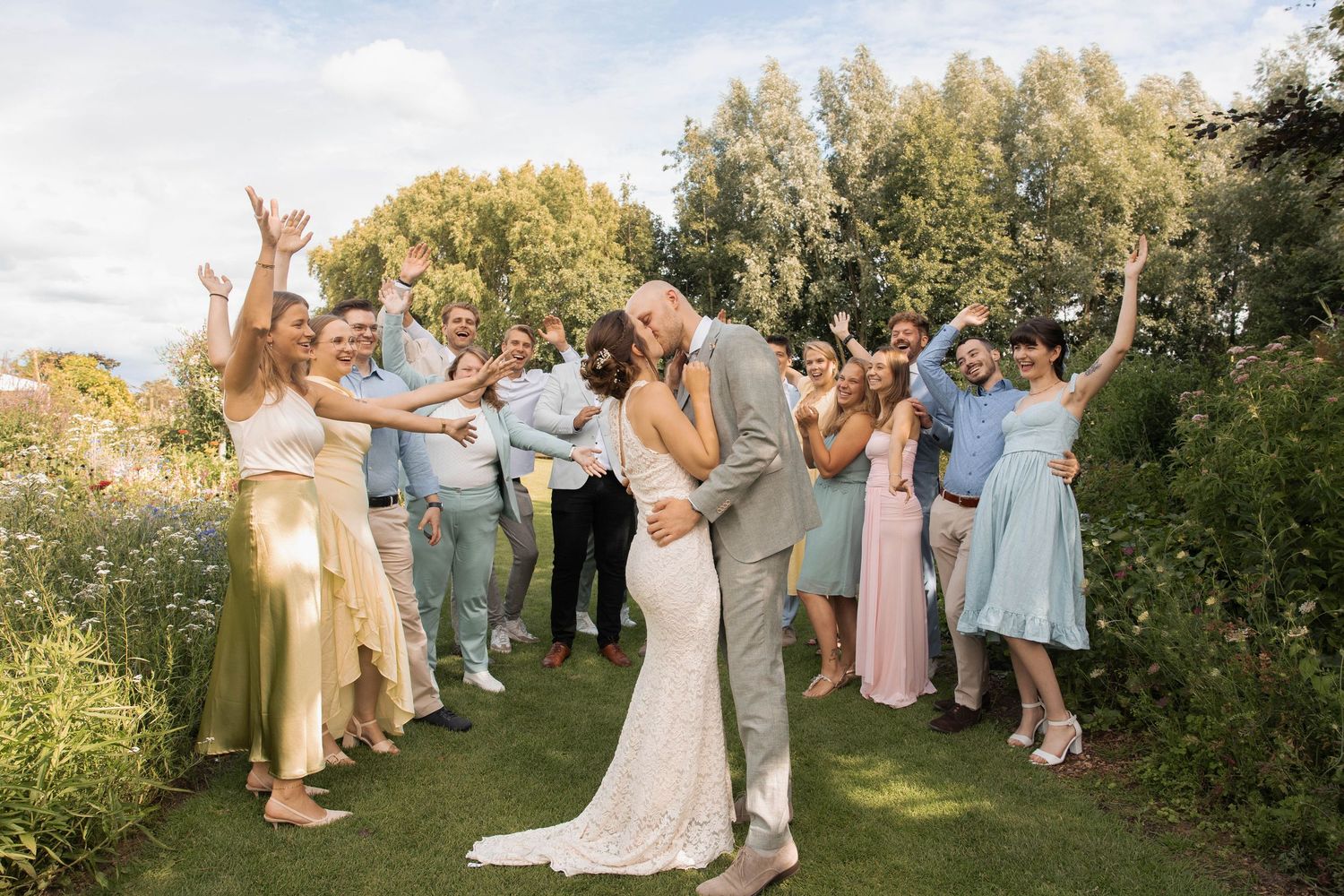 Bride and groom share a kiss surrounded by cheering wedding guests in a lush garden setting.