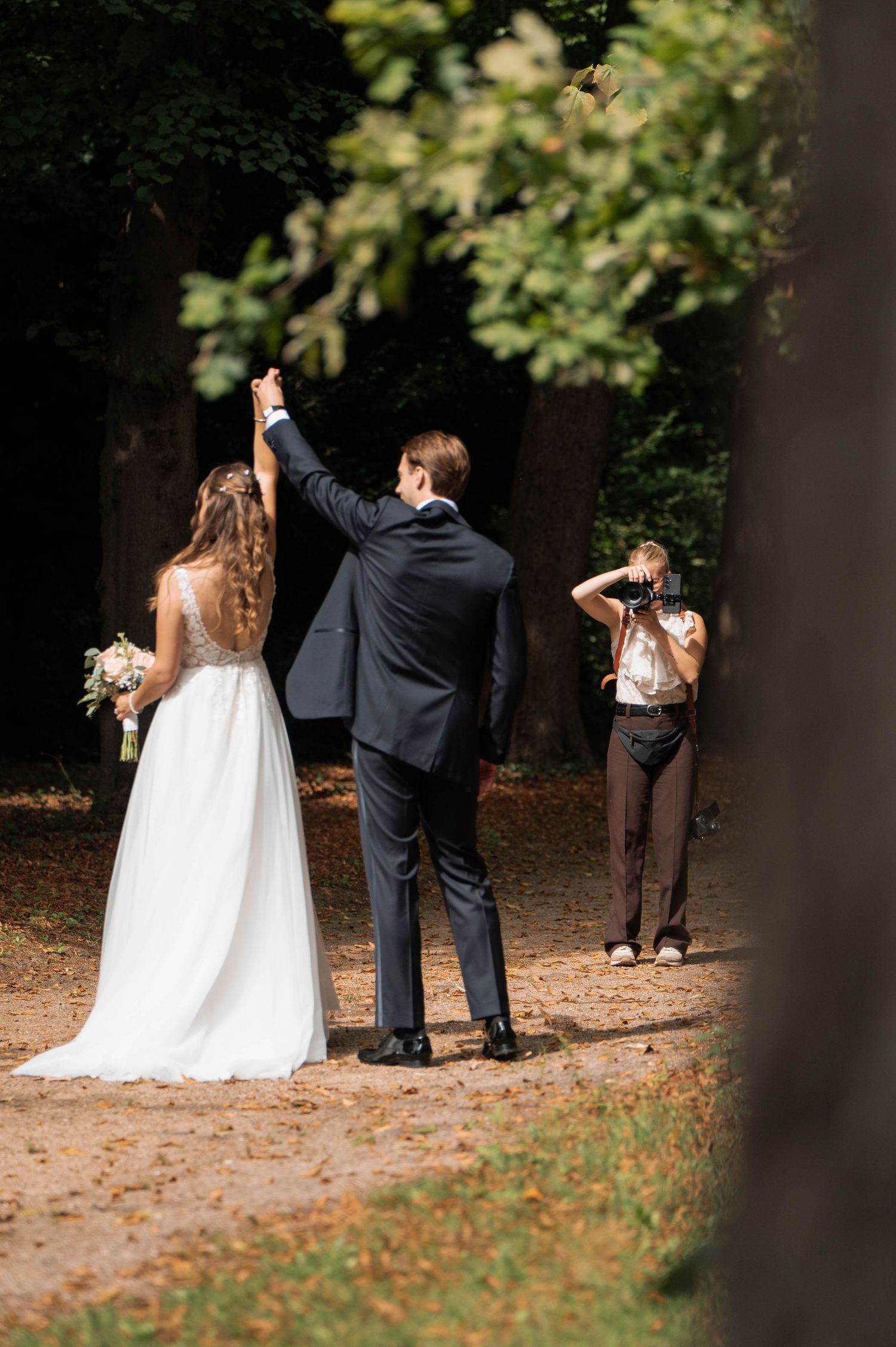 Bride and groom holding hands with photographer capturing their wedding portrait in a lush green park setting.