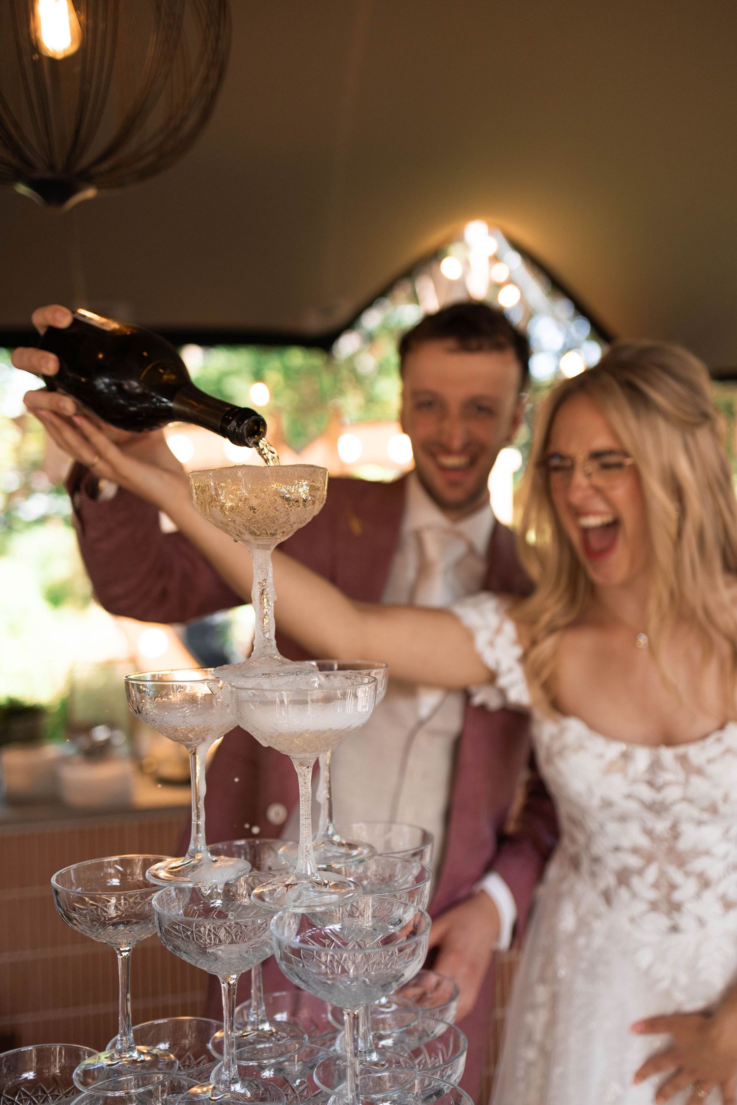 Laughing bride and groom pour champagne over a glass tower of coupe glasses at a joyful wedding celebration.