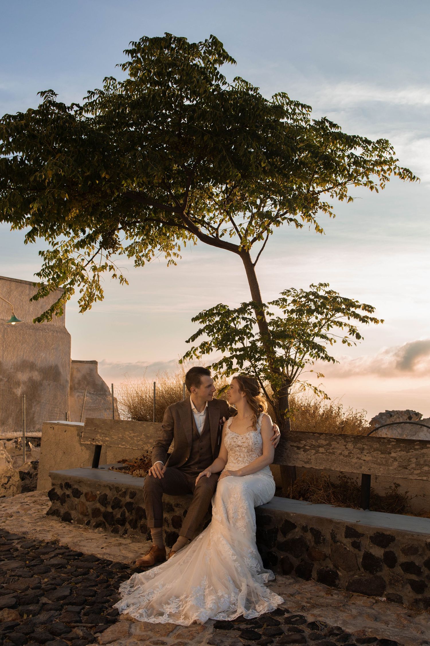 Bride in a lace wedding gown and groom in a suit sit close on a stone bench under a tree at sunset, with rustic walls behind them.