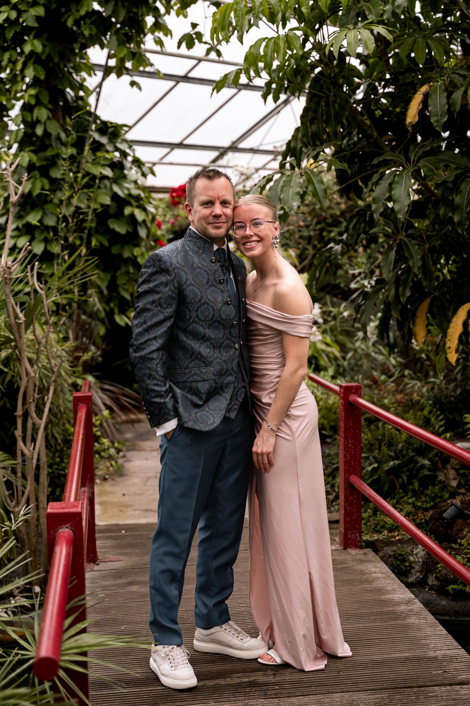 Couple posing on wooden bridge in lush greenhouse, woman in pink off-shoulder gown, man in dark patterned vest.