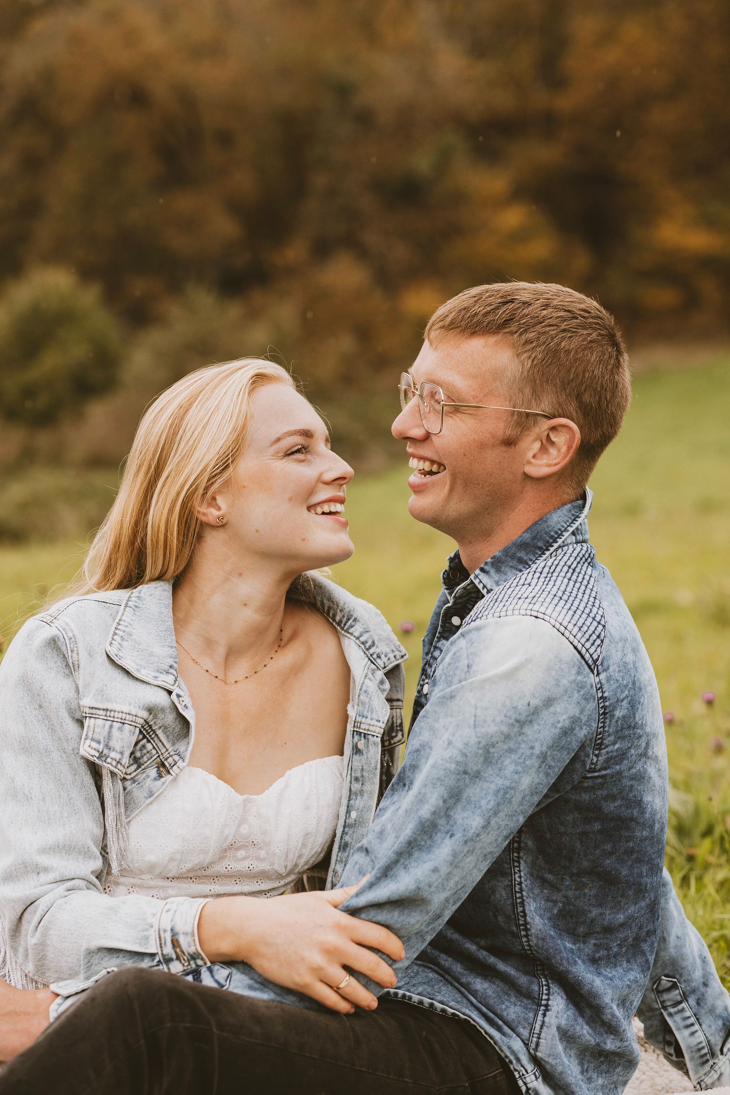 A couple shares joyful laughter while sitting together outdoors in autumn wearing matching denim jackets.