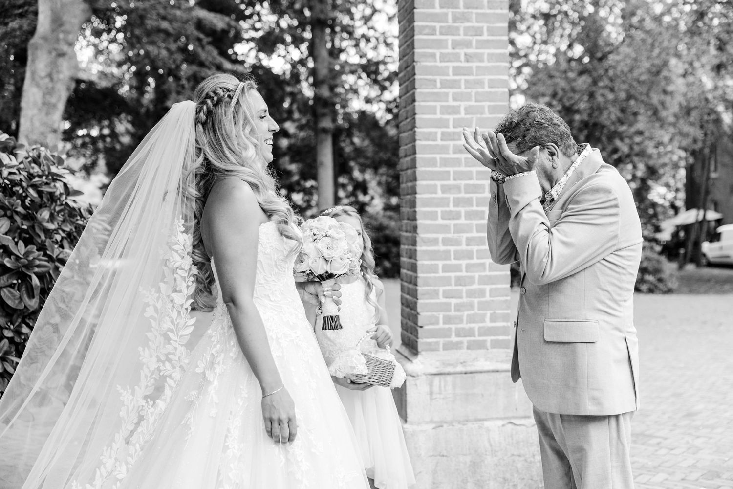 Black and white photo of emotional groom covering face as bride in wedding gown and veil stands before him.