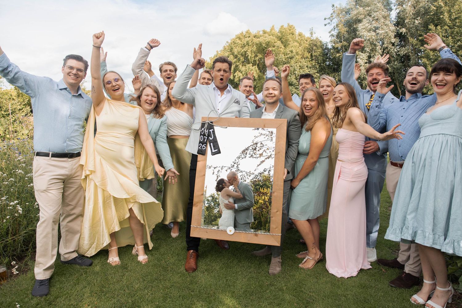 Wedding party celebrates outdoors holding a framed photo, guests cheering with raised arms on a sunny day.