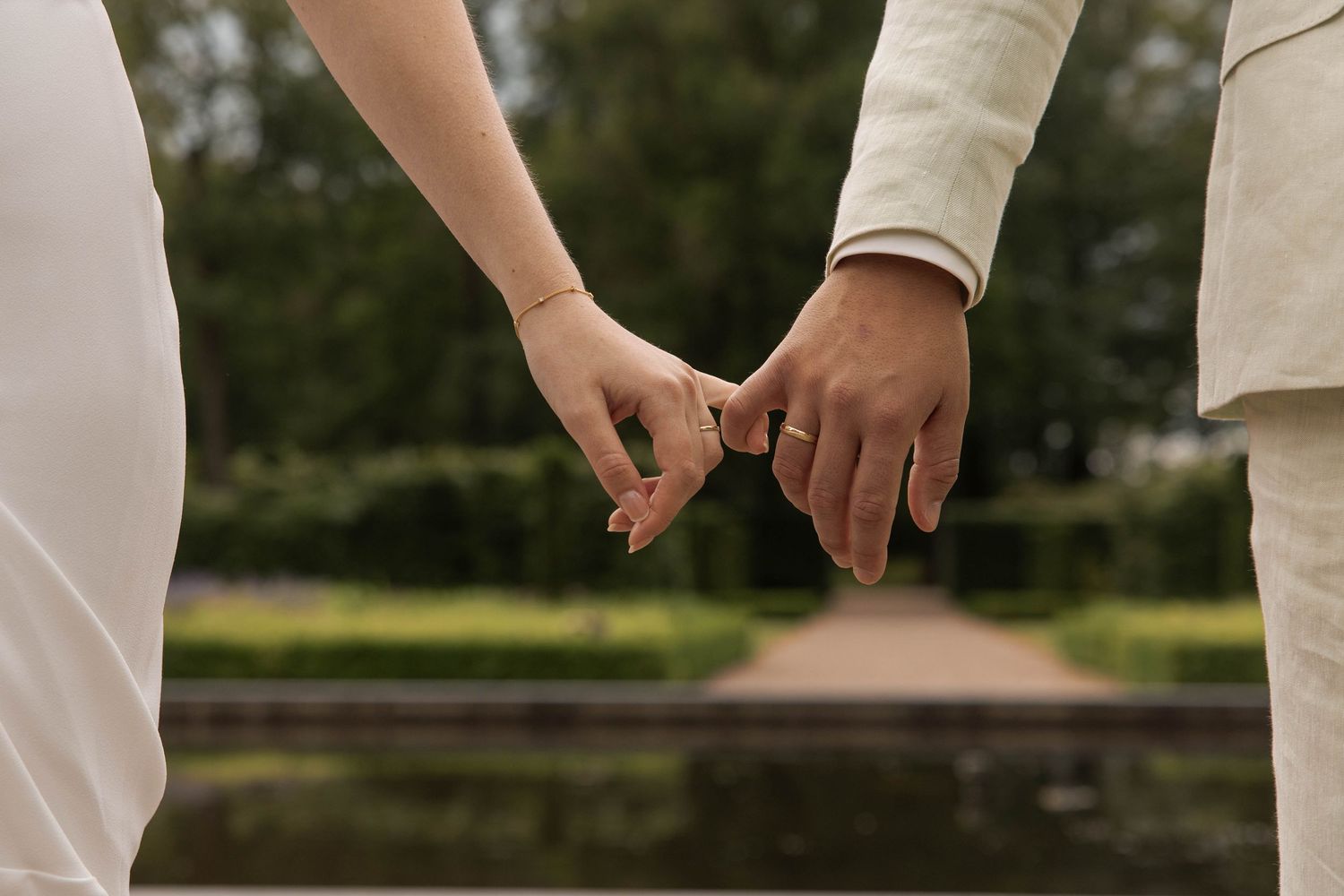 Couple holding hands gently in a garden setting, wearing wedding attire, symbolizing love and commitment.