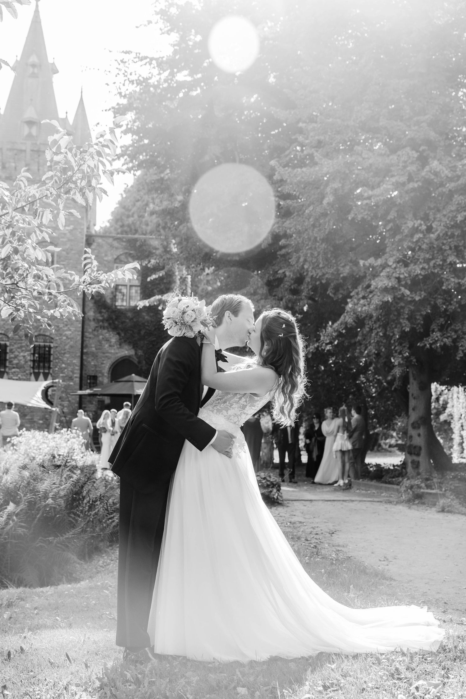 Black and white photo of a bride and groom sharing a kiss outdoors near a castle with sunlight flare.