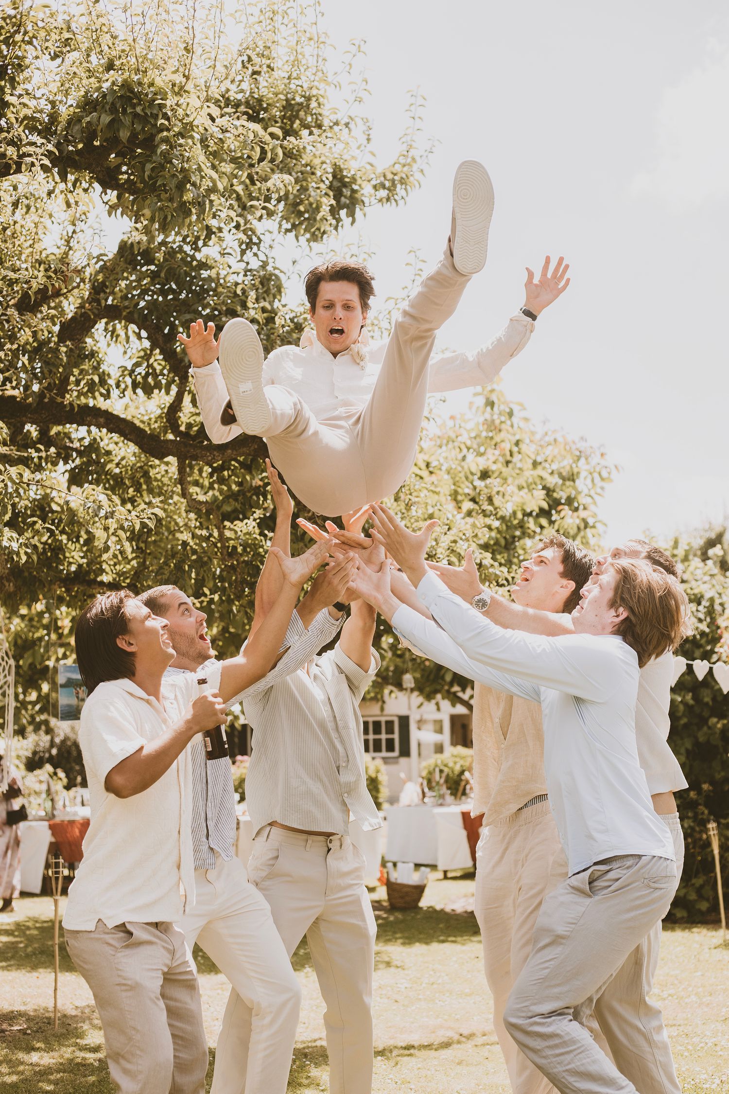 Groomsmen in light colored suits playfully toss groom in the air during outdoor wedding celebration in sunlit garden setting.