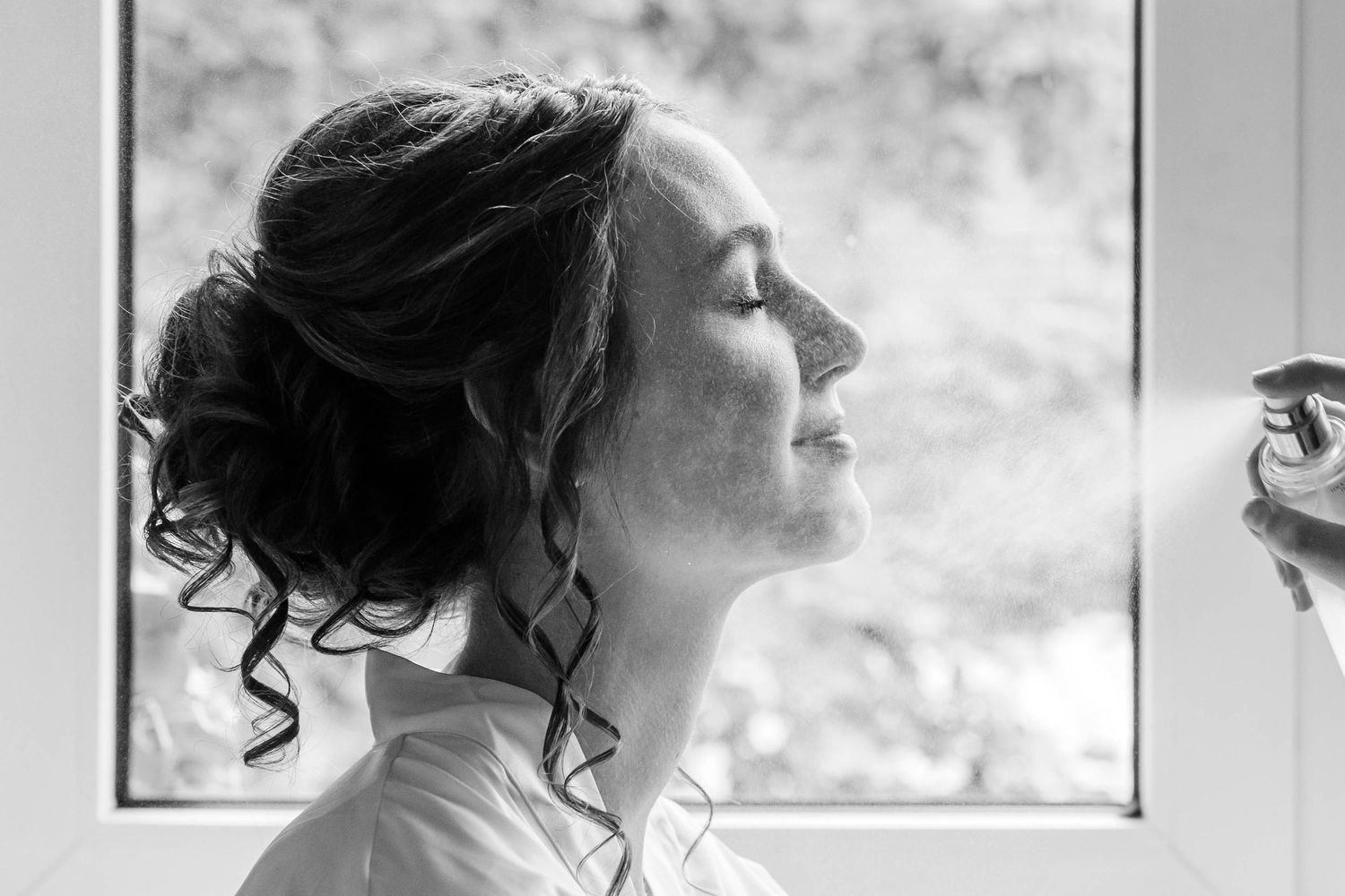 Black and white photo of a bride spraying perfume with eyes closed near a bright window.