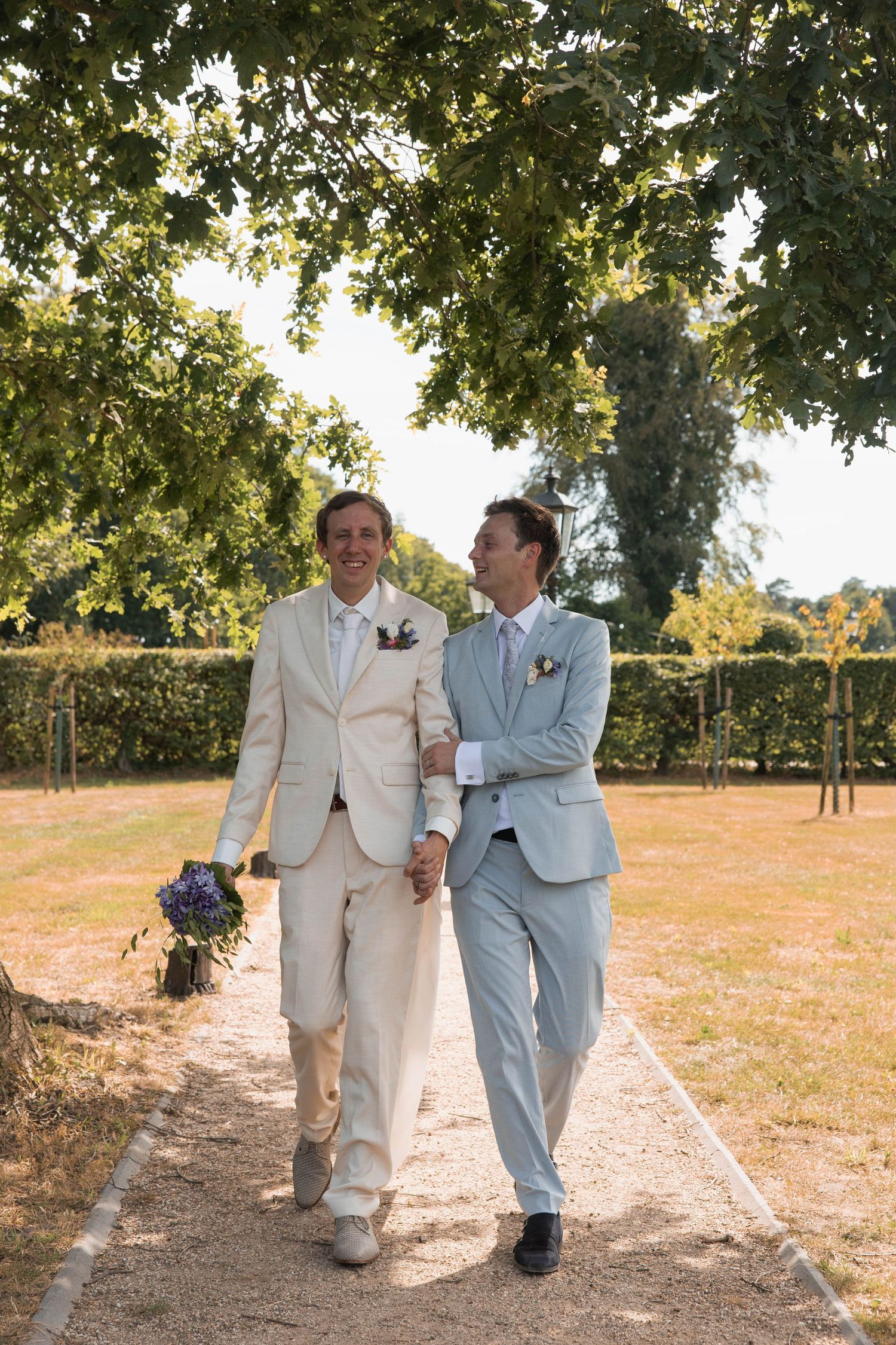 Two grooms in suits walk hand in hand through a sunny garden path on their wedding day.