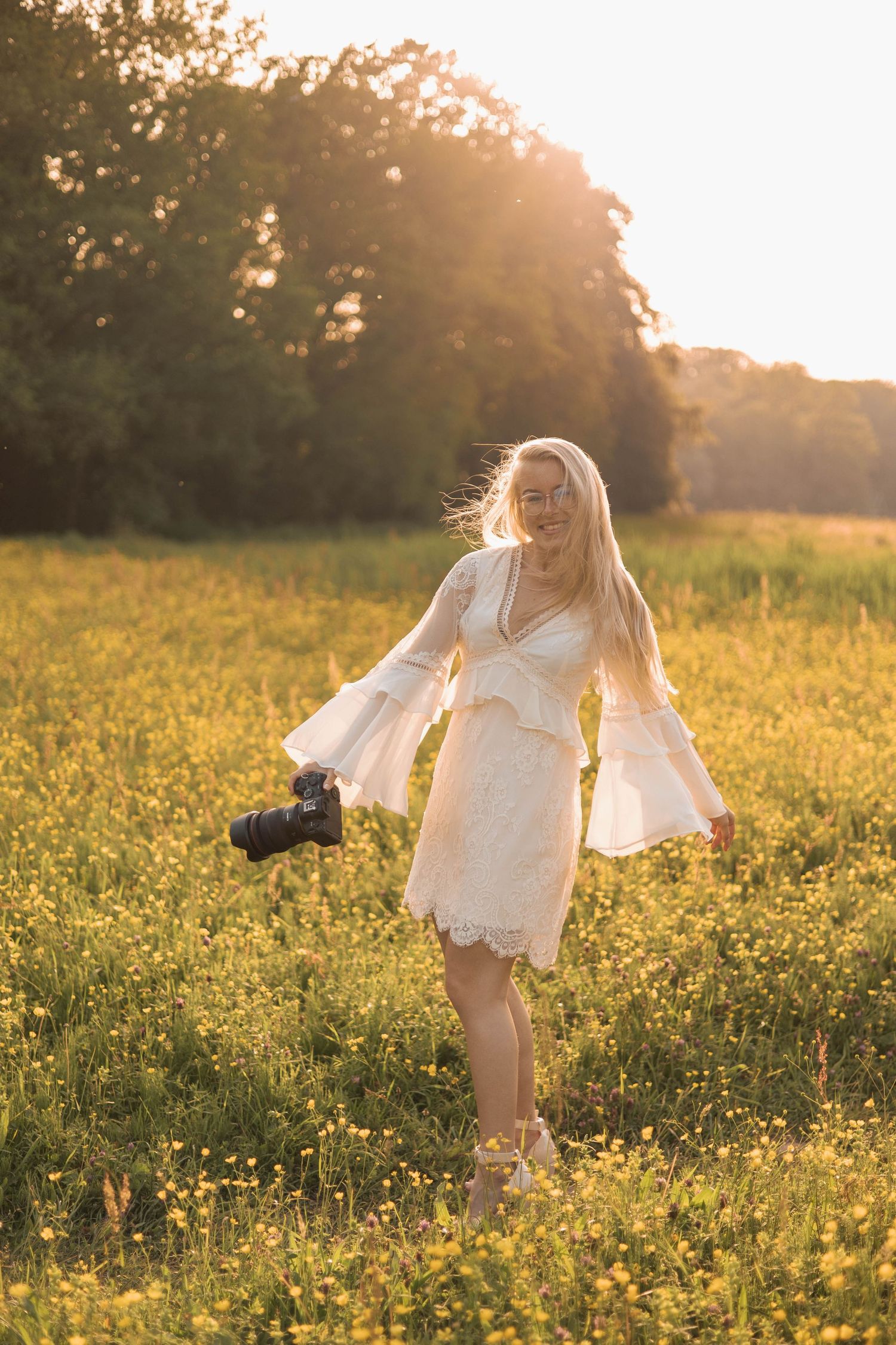 Blonde woman in white lace dress holding camera, walking through golden flower field at sunset.