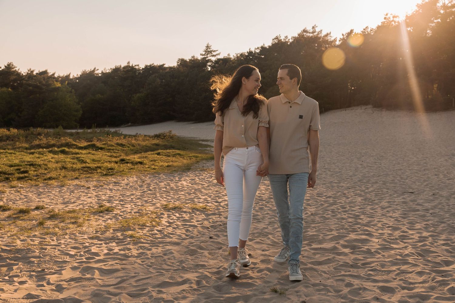 Couple walking hand in hand on sandy dunes at golden sunset with pine trees in background.