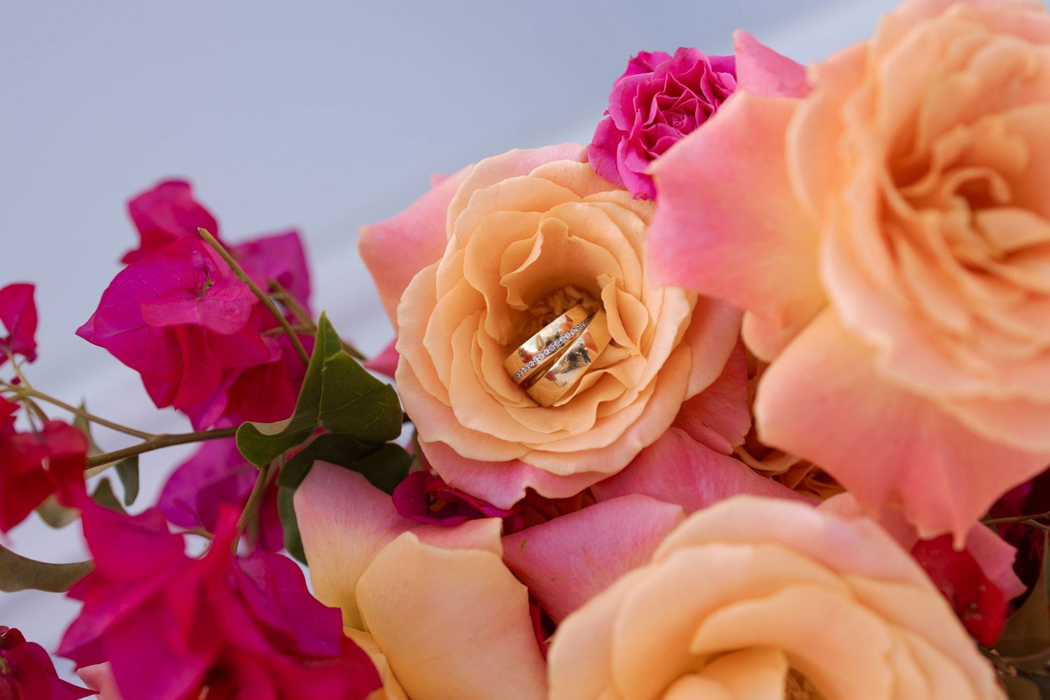 Two gold wedding bands resting in the center of a peach rose surrounded by pink roses and magenta bougainvillea blooms.