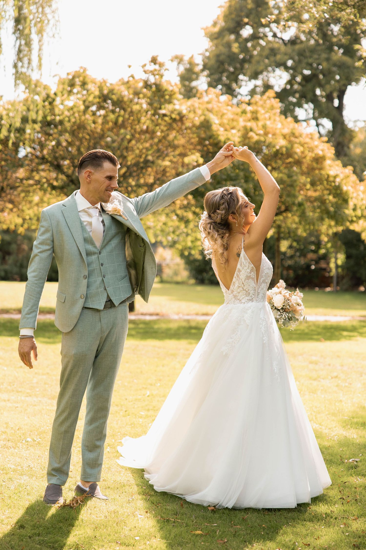 Bride and groom dancing outdoors in a sunlit garden, groom in sage suit twirling bride in white lace wedding gown.