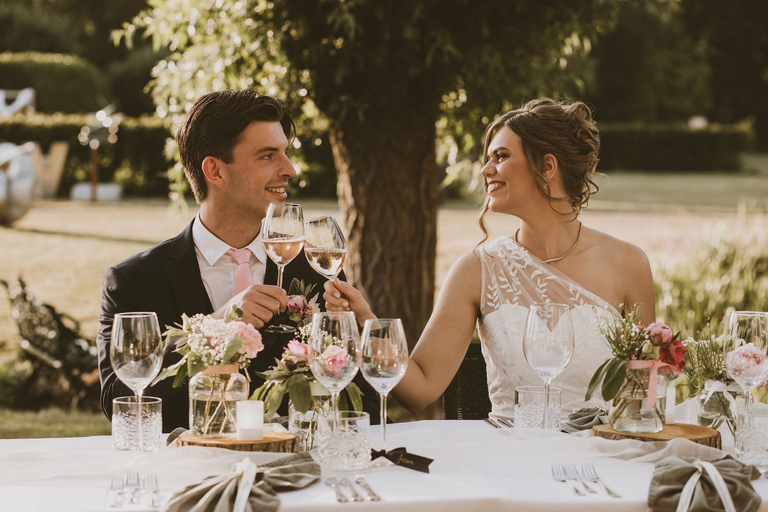 A couple toasts with wine glasses at an elegant outdoor wedding reception table decorated with floral centerpieces.
