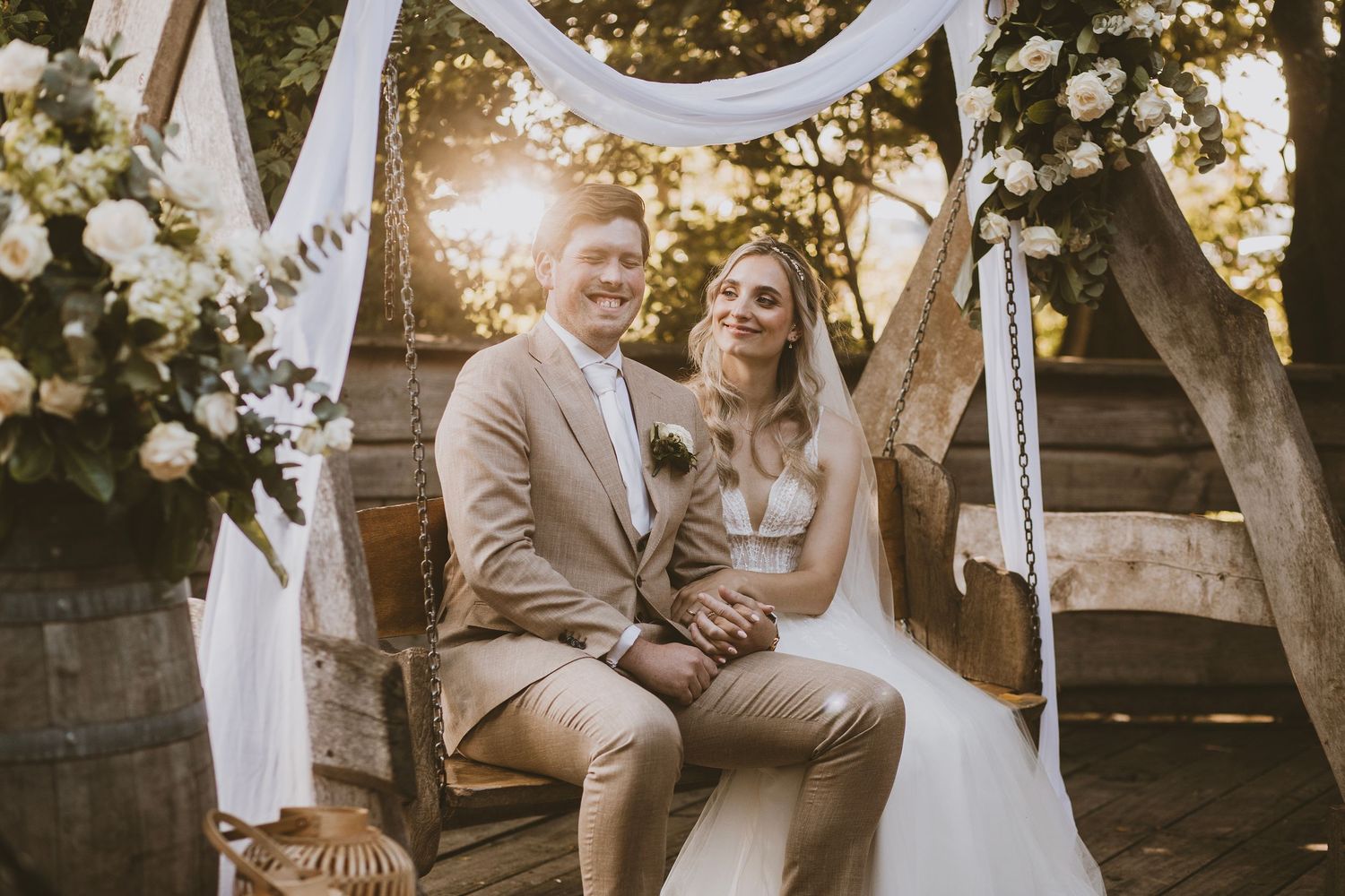 A romantic outdoor wedding ceremony setup with white drapes and floral decorations during golden hour sunset.
