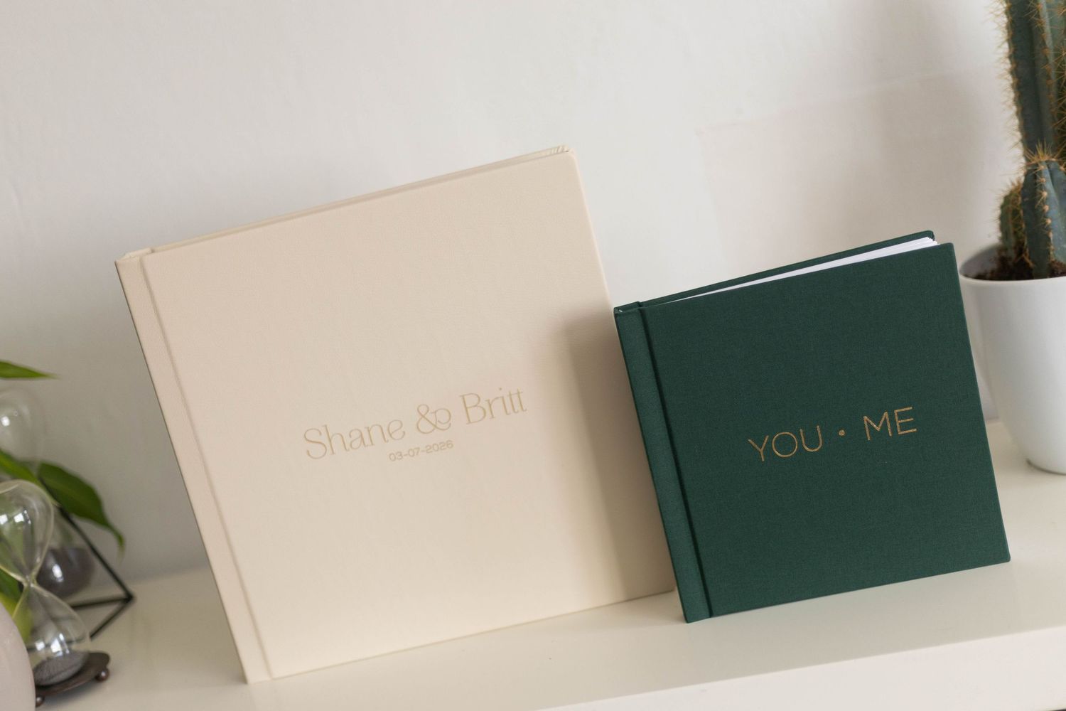 Two elegant wedding photo albums, one cream with gold text and one dark green, displayed on a white shelf.