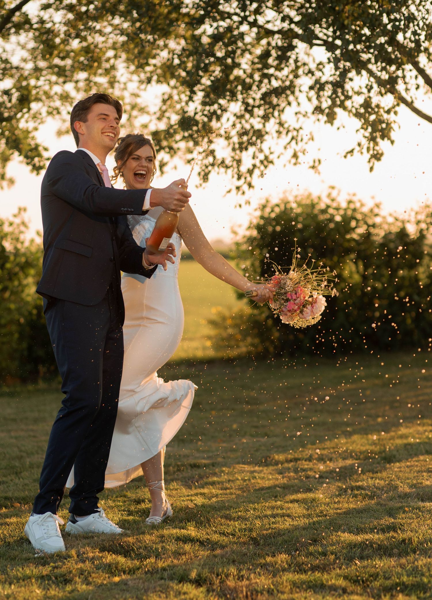Joyful newlyweds popping champagne outdoors at golden hour, bride holding pink bouquet in white gown, groom in navy suit.