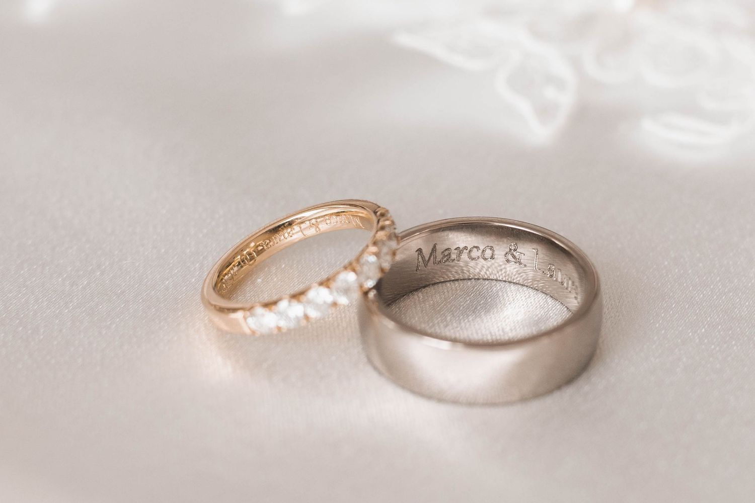 Two wedding rings, one gold with diamonds and one silver engraved band reading Marco, rest on a white surface.